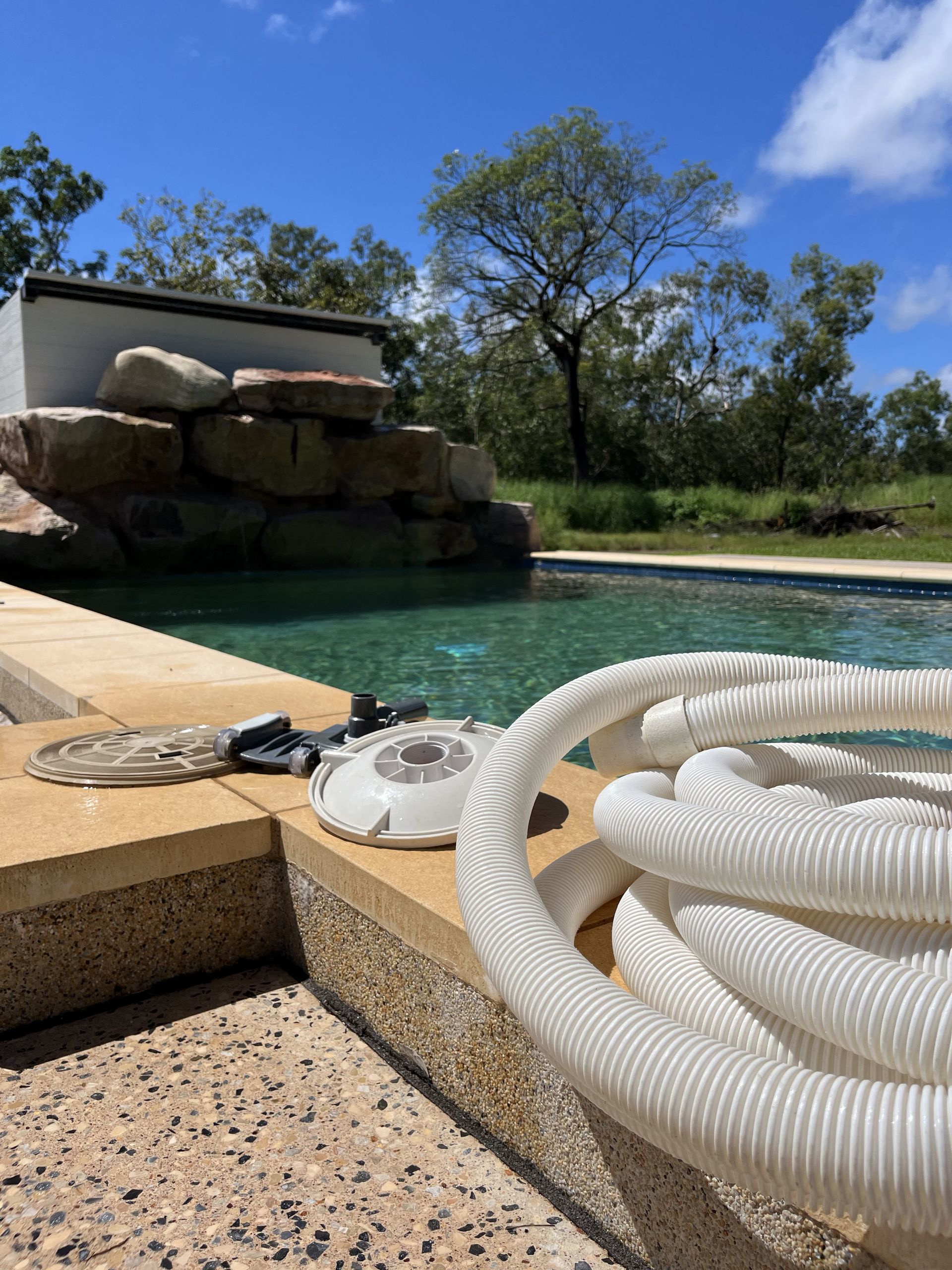 Pool cleaning equipment, including a coiled white hose and suction head, sits on the edge of a backyard pool  — Friendly Pool & Spa Care in Yarrawonga, NT 