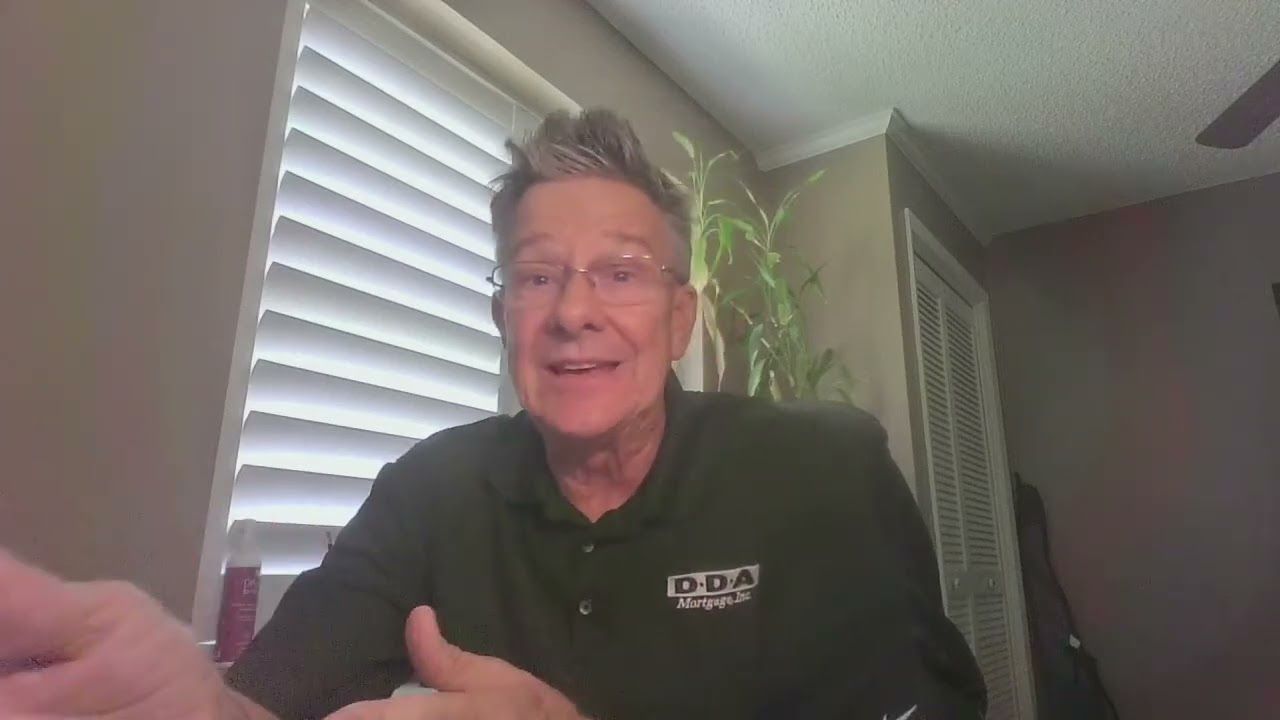 Person speaking on a webcam in a home office, wearing a black shirt near a window with blinds and a plant.