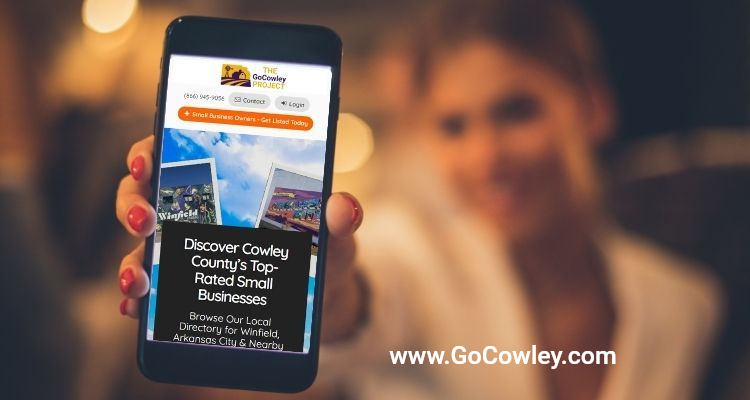 A hand holds a smartphone displaying a business directory website for Cowley County against a blurred background.