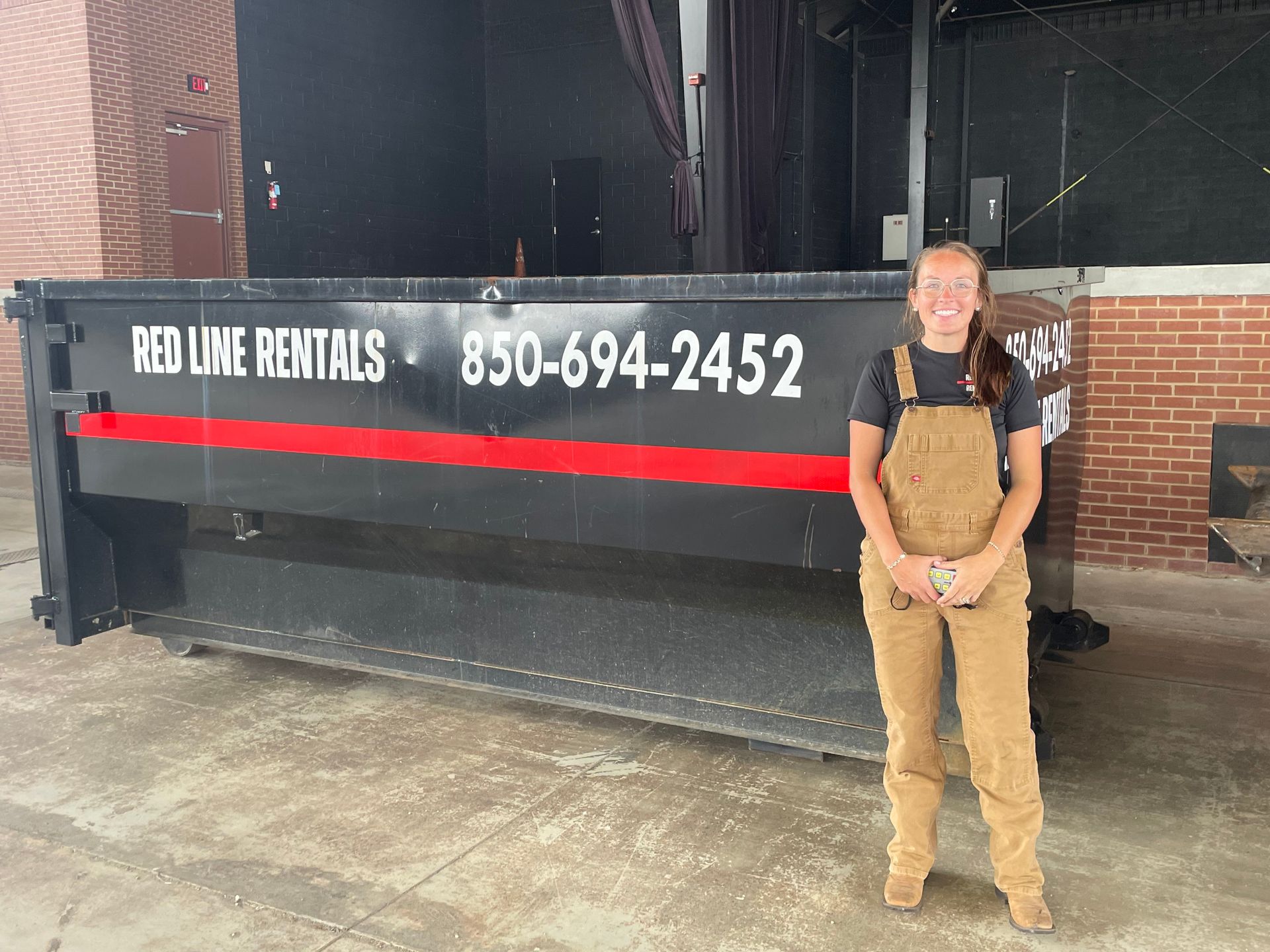 Woman in overalls standing next to a Red Line Rentals dumpster inside a building.