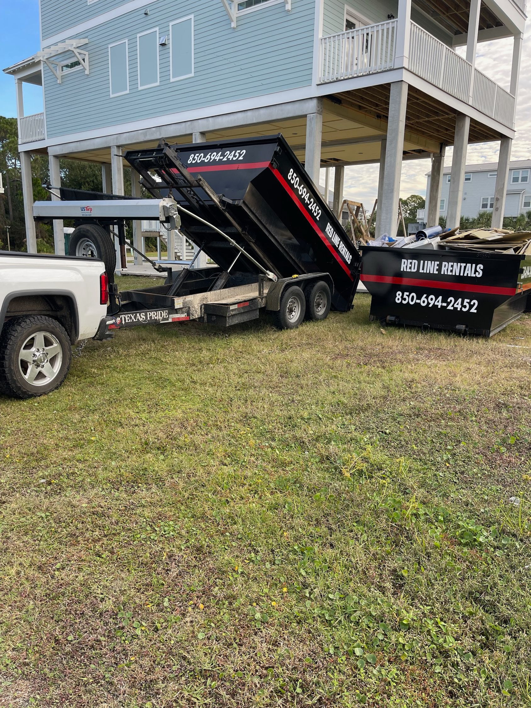 A white pickup truck with an elevated black dump trailer parked on a grassy lot next to a stilted house.