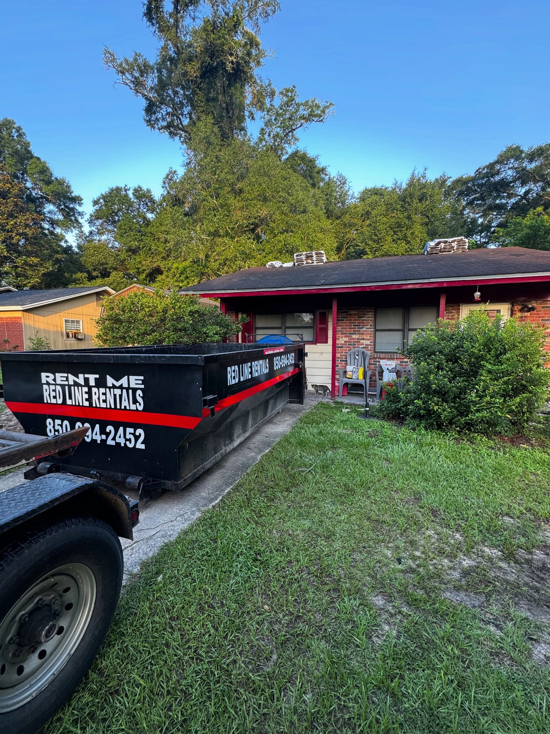 Black and red dumpster trailer in front of a brick house with overgrown yard, trees and blue sky.