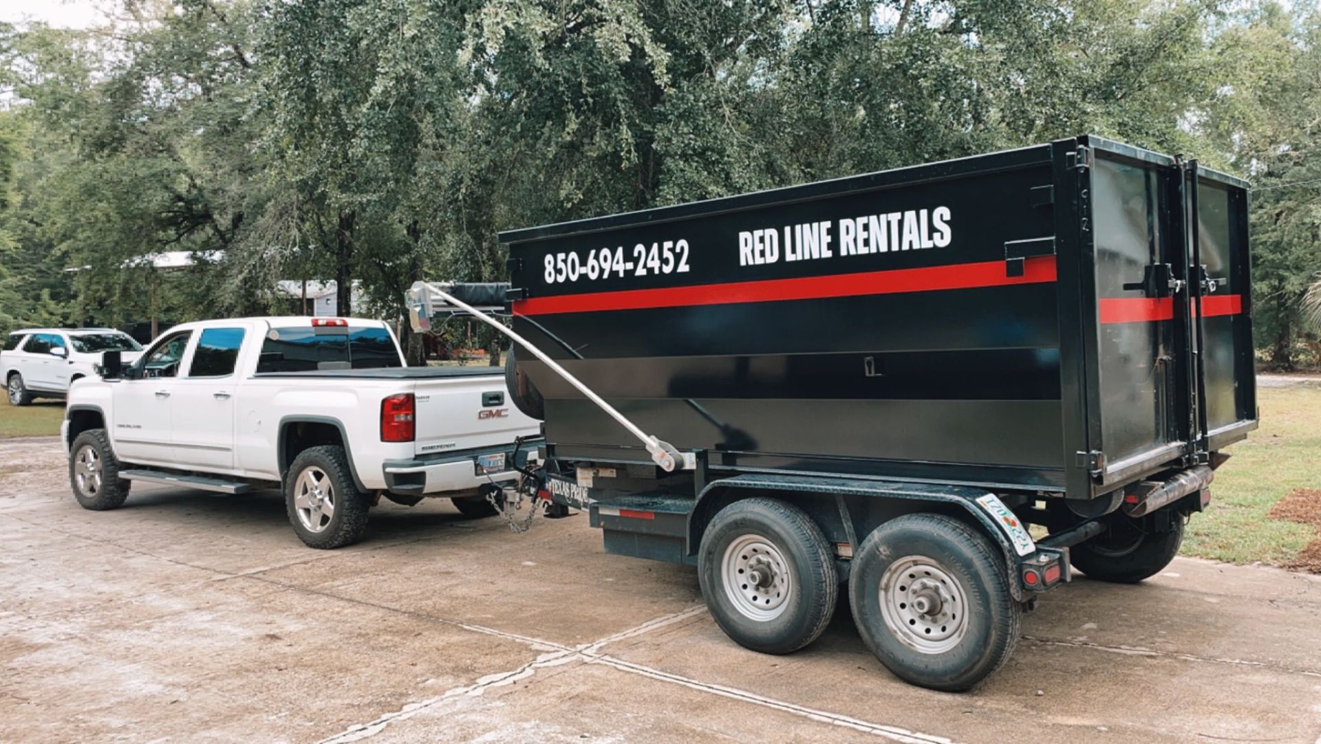 A white pickup truck towing a black Red Line Rentals dumpster trailer parked on a gravel surface outdoors.