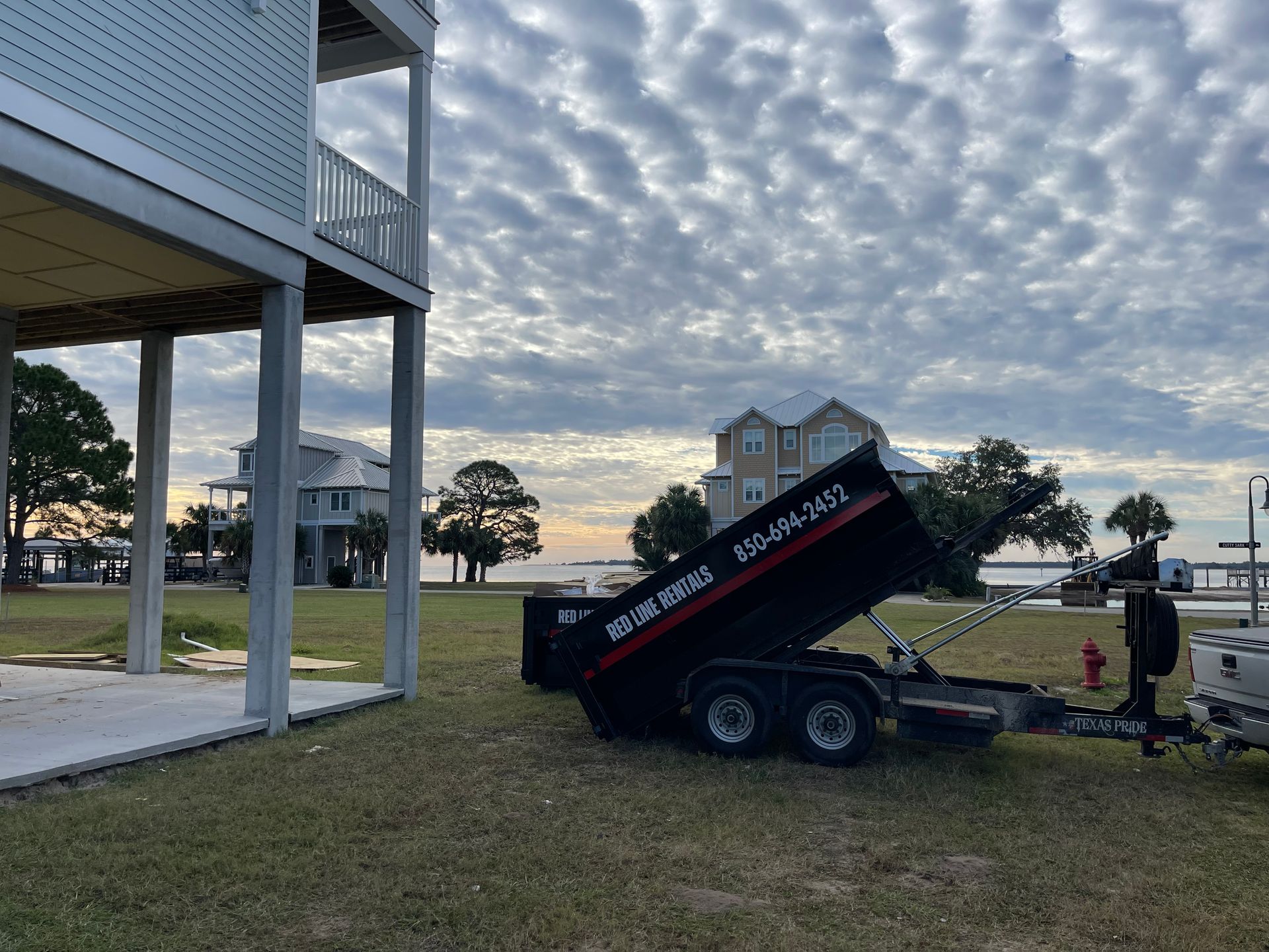 A black dump trailer next to a house under a cloudy sky near a body of water.