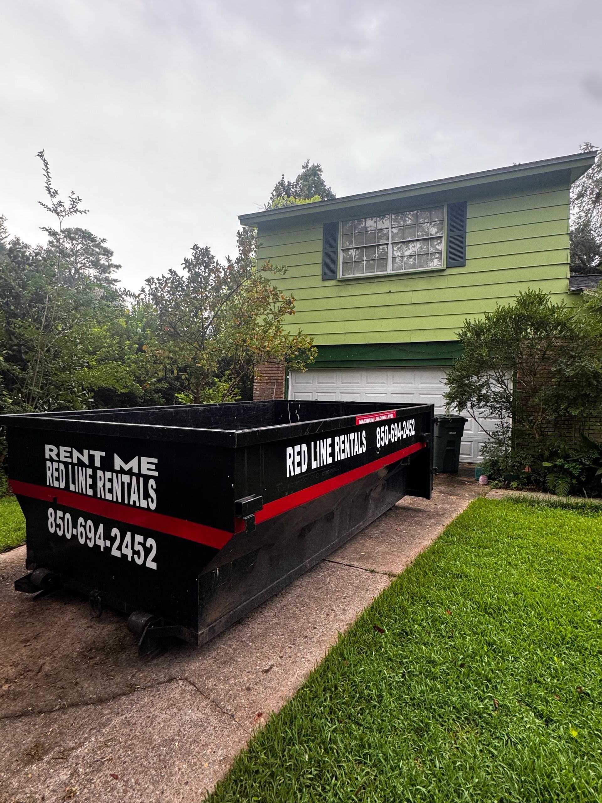 Black dumpster in front of a green house with a closed garage door.  The dumpster has red and white text.