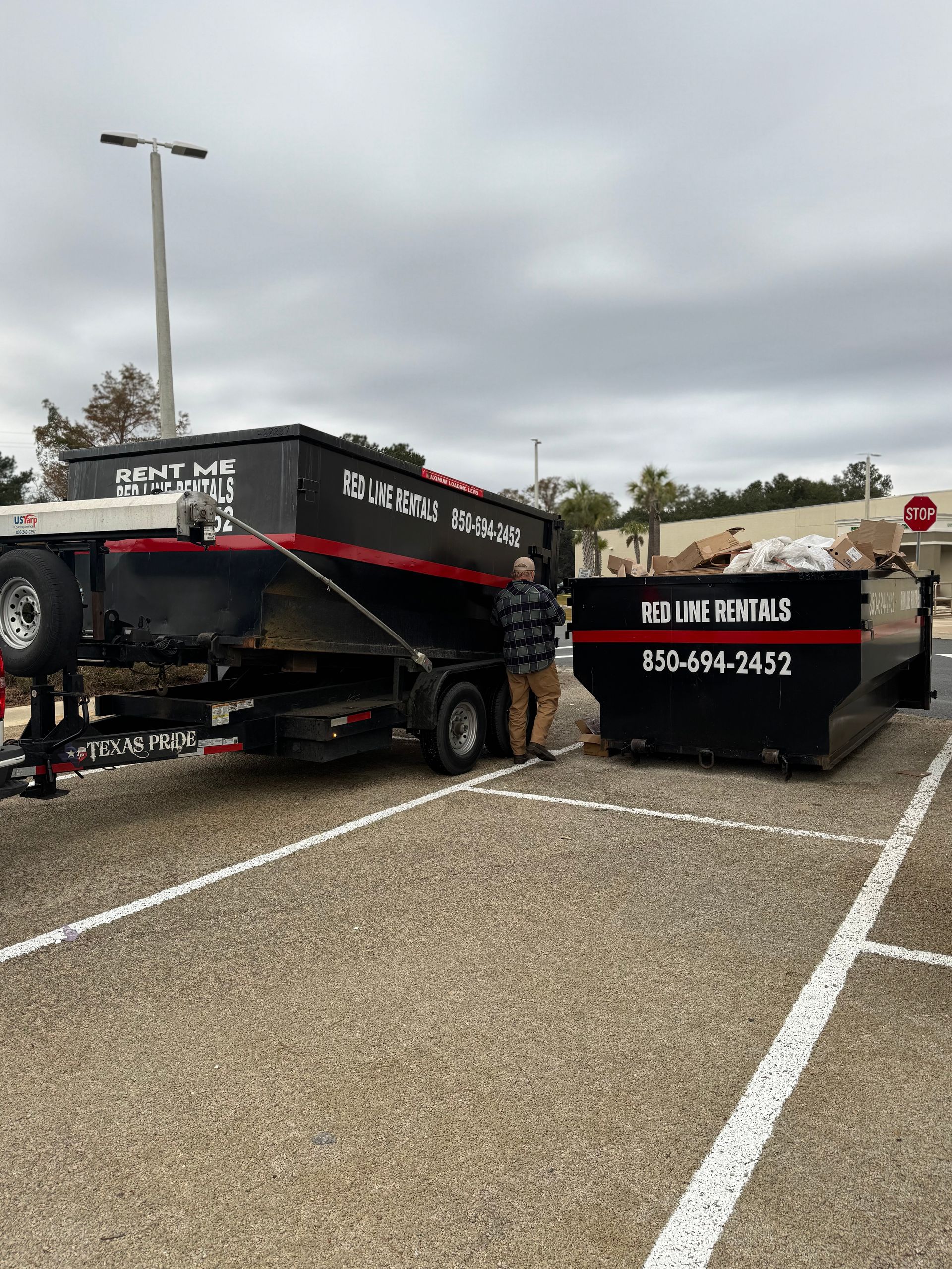 Two black roll-off dumpsters on a gravel lot; one on a trailer. A man stands between them under a cloudy sky.