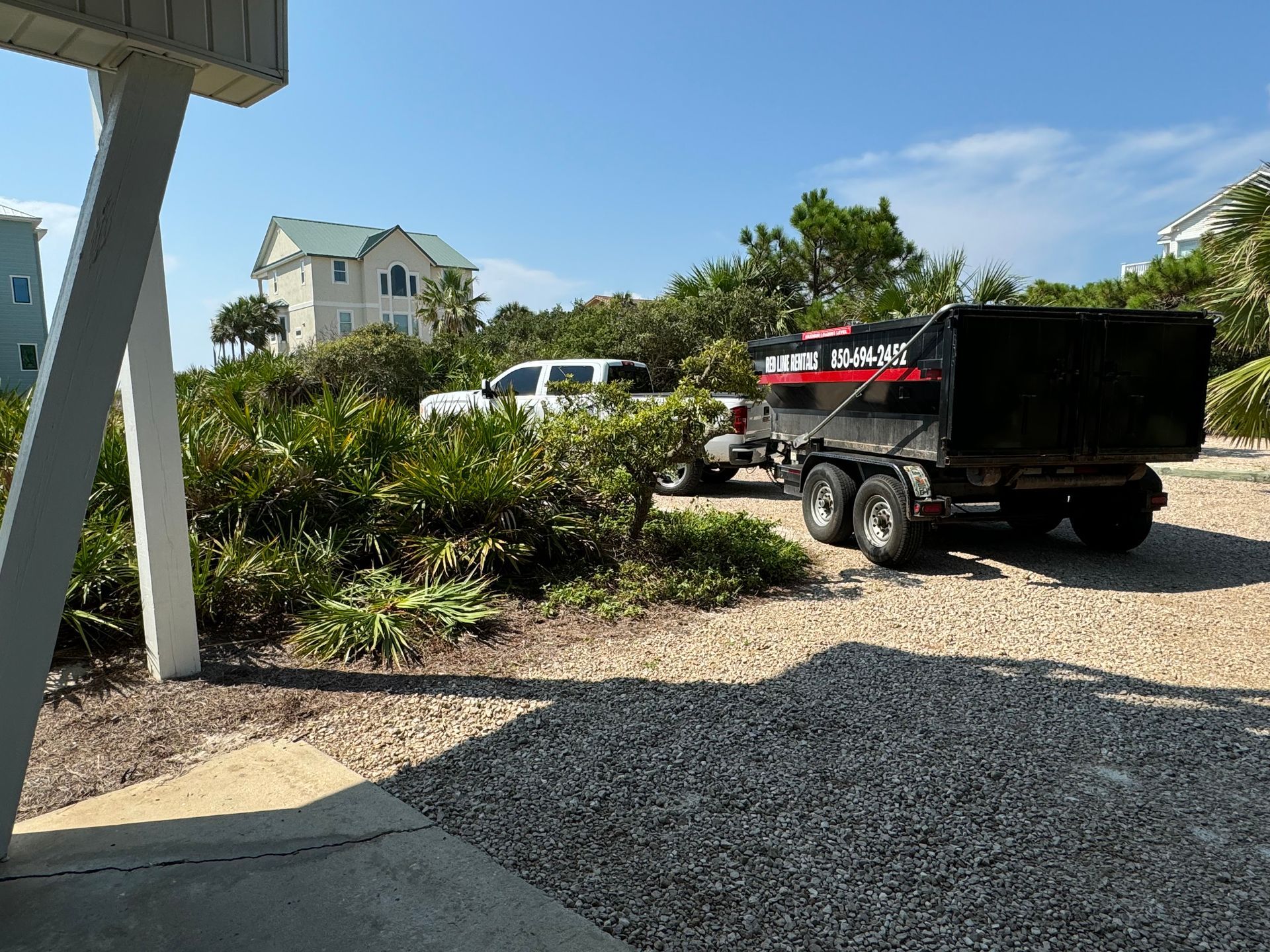 White truck towing a black dump trailer on a gravel driveway, with houses and greenery under a blue sky.