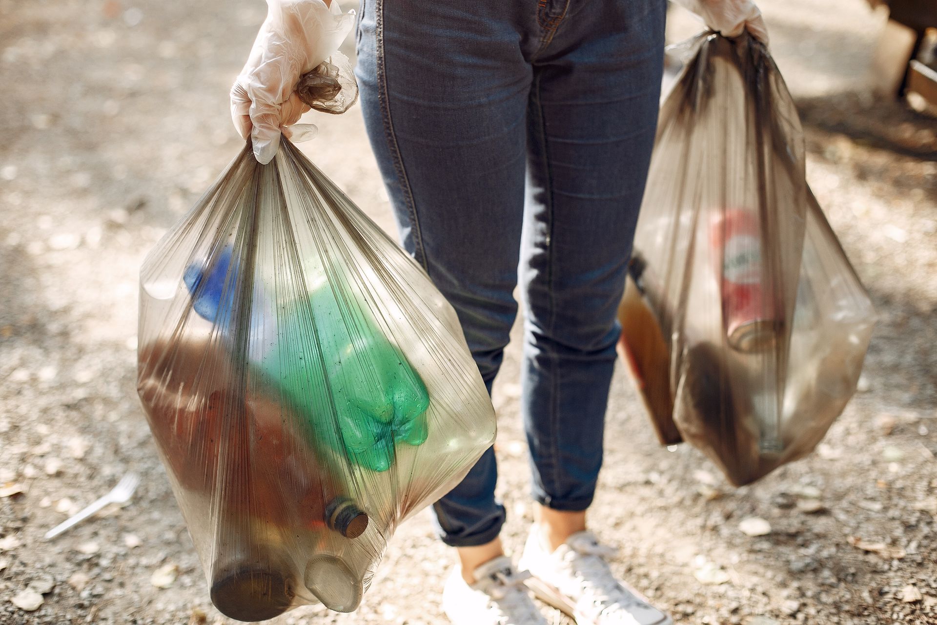 A woman is carrying two bags of trash on a dirt road.