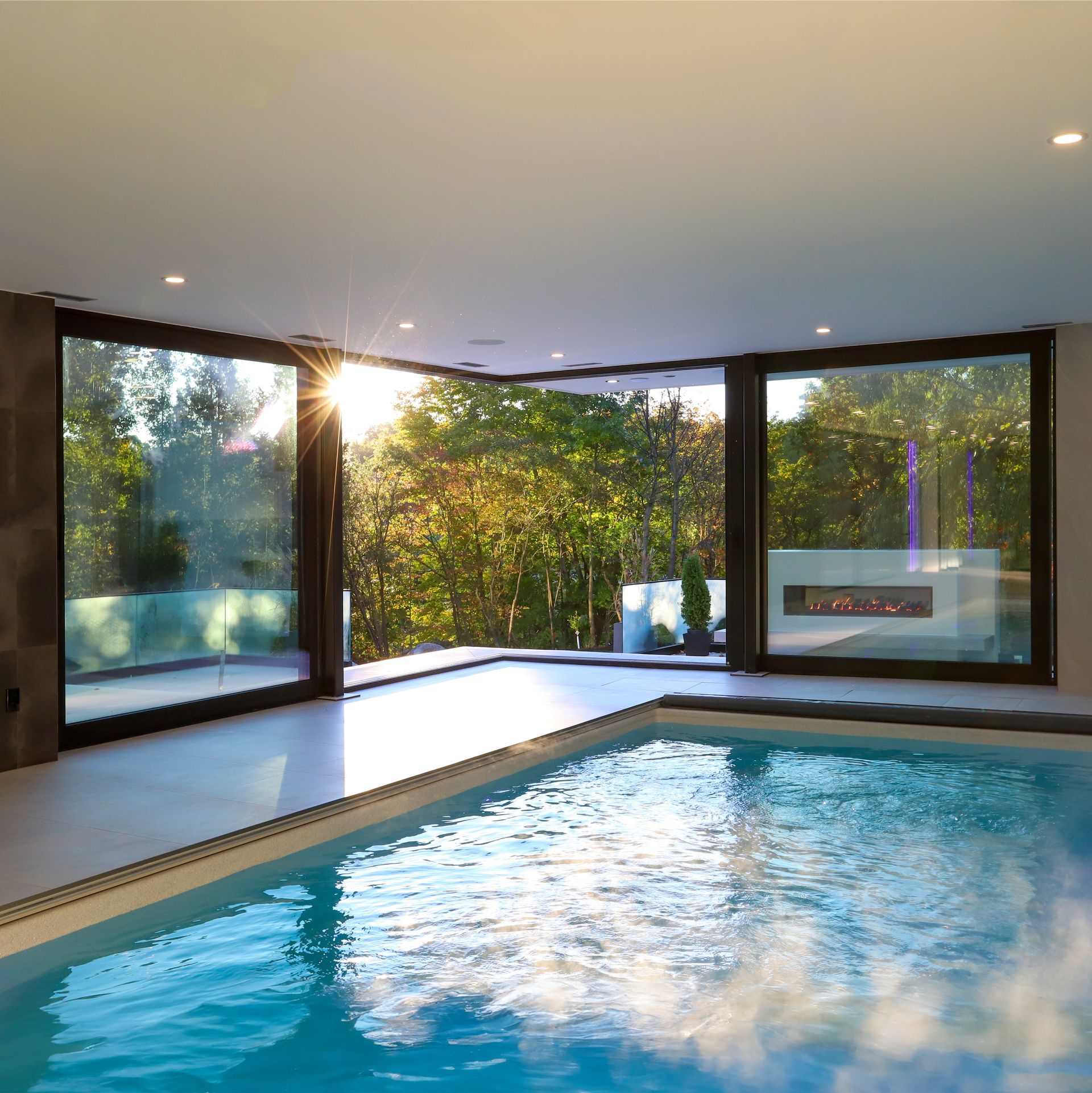 Indoor swimming pool with large windows, bright sunlight, and a view of trees.
