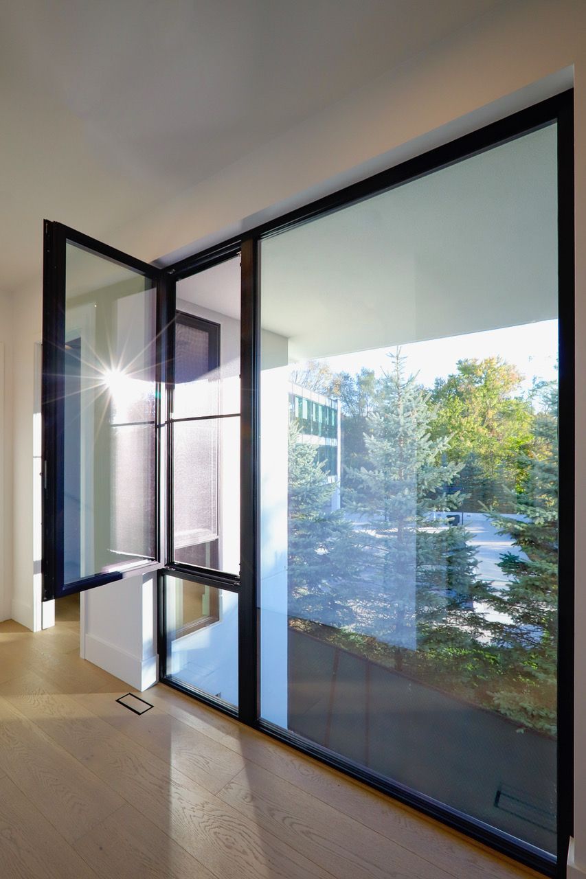 Black-framed glass door, one section open, overlooking trees and a building; interior with wood floor.