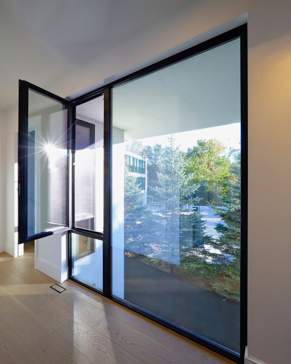 Large black-framed window partially open; view of trees, sunlight reflecting on glass; modern interior.