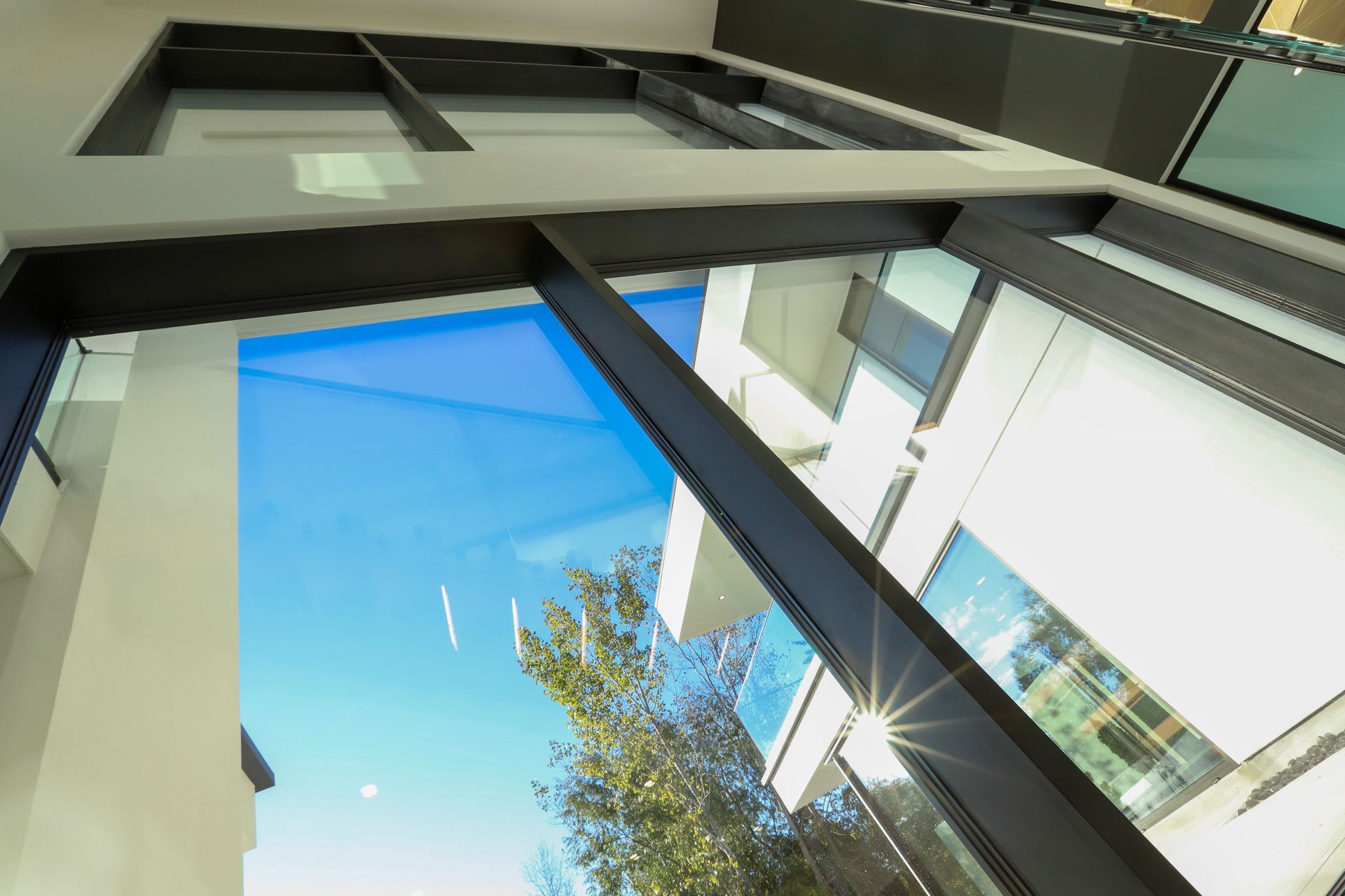 Looking up through modern windows, blue sky, and a glimpse of a white building and green tree.
