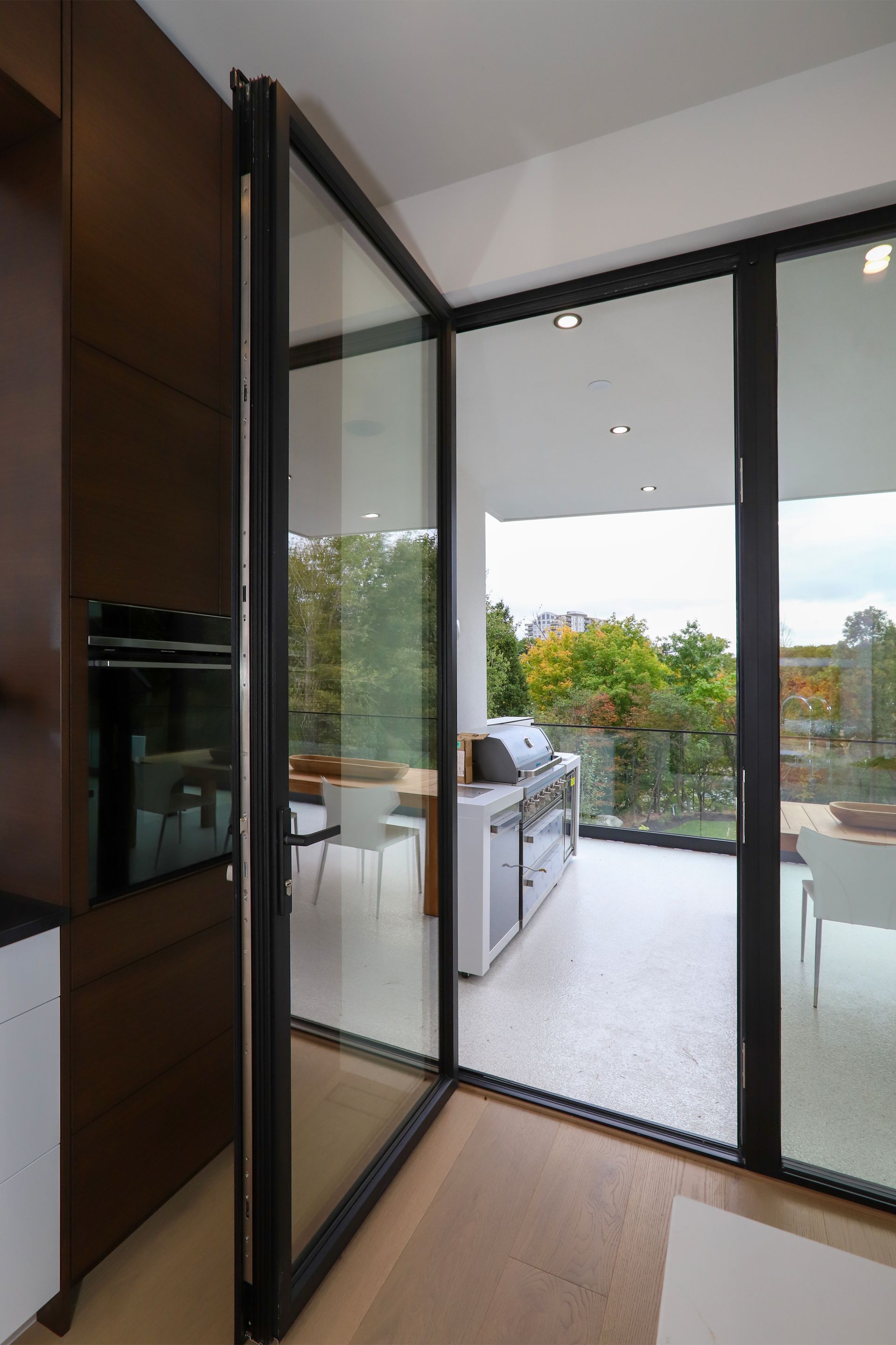 Bifold glass doors open to a patio with a grill. Interior kitchen with dark cabinets.