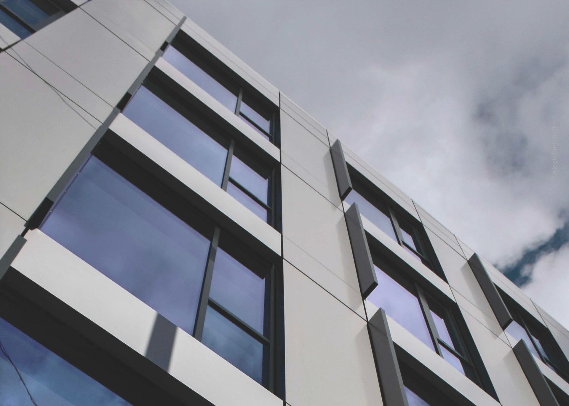 Modern building facade with white panels, dark-framed windows, and a cloudy sky.