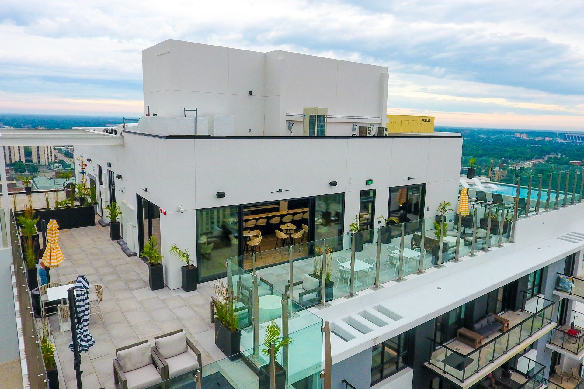 Rooftop bar with outdoor seating, glass railings, and a city view on a cloudy day.