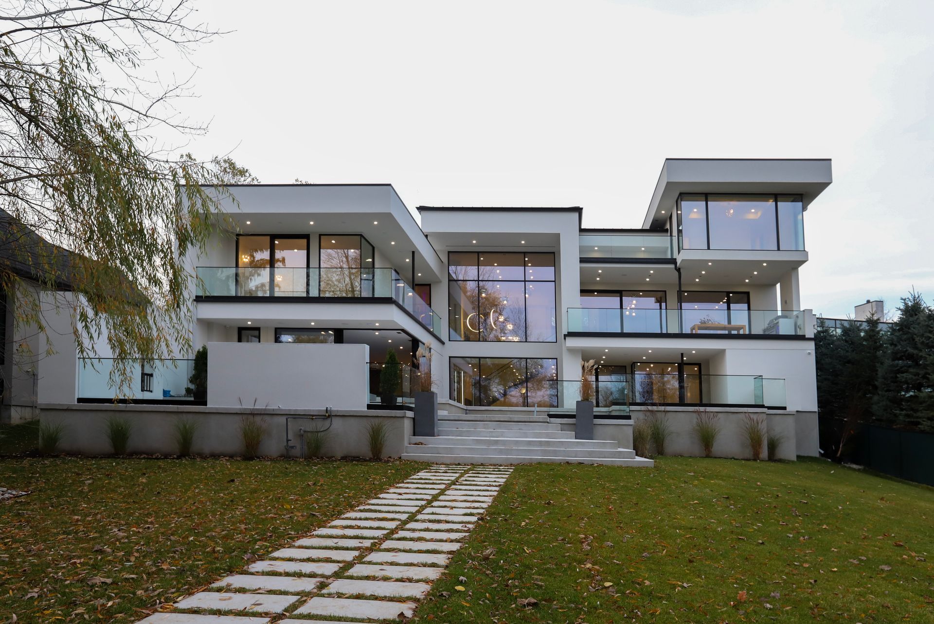 Modern white multi-story house with glass windows and balconies. Stone path leads from the lawn.