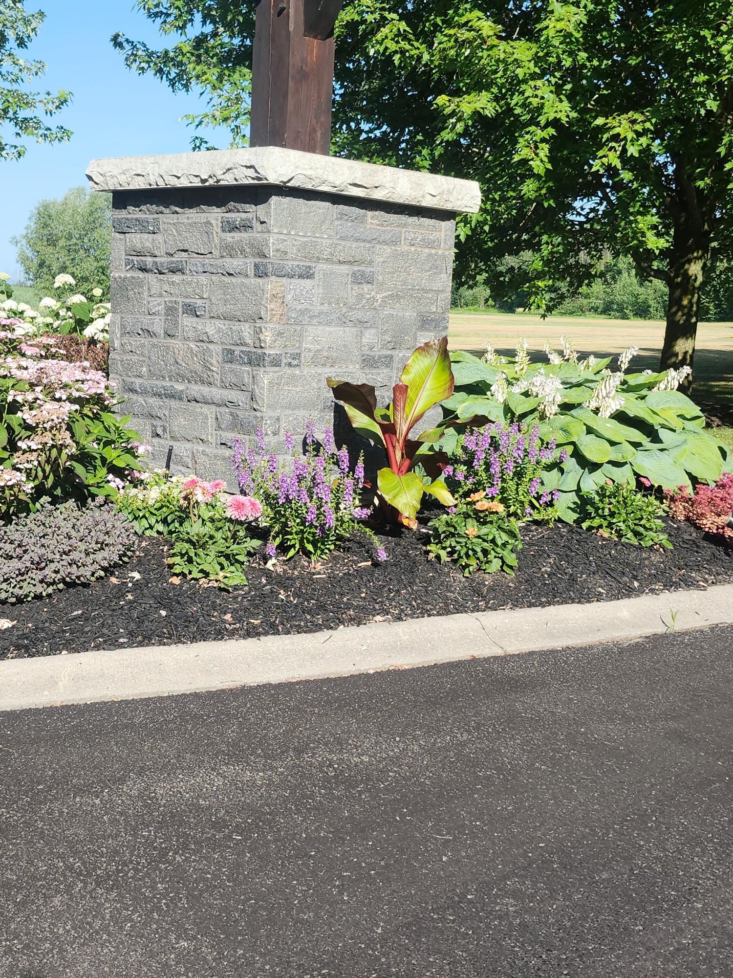A brick house porch with a white column, surrounded by green shrubs and hostas in a mulch bed next to a grass lawn.