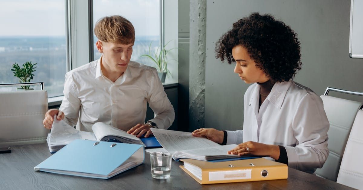 Two people in white shirts reviewing documents at a desk by a window.