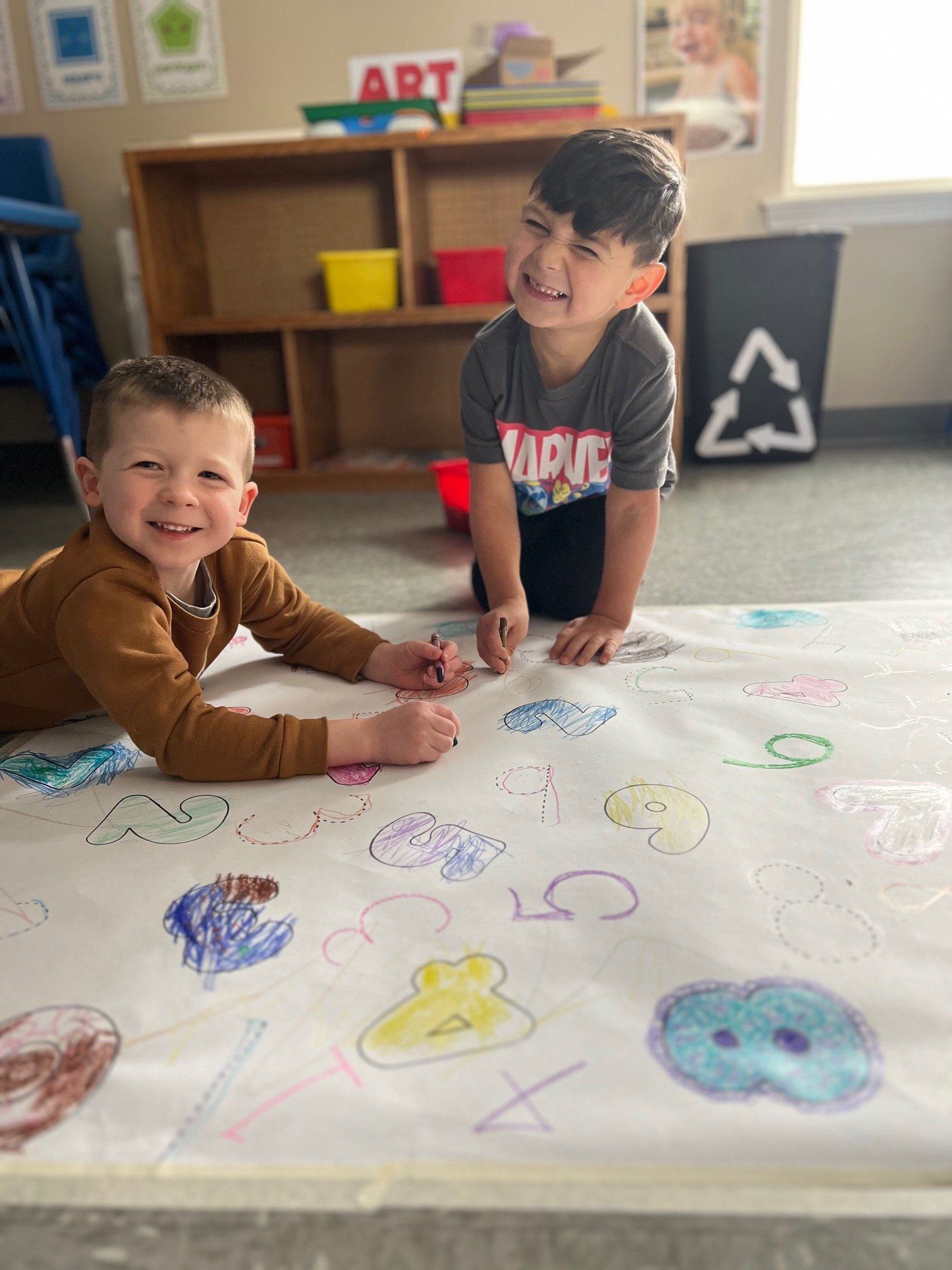 Two young boys are laying on the floor drawing on a large piece of paper.