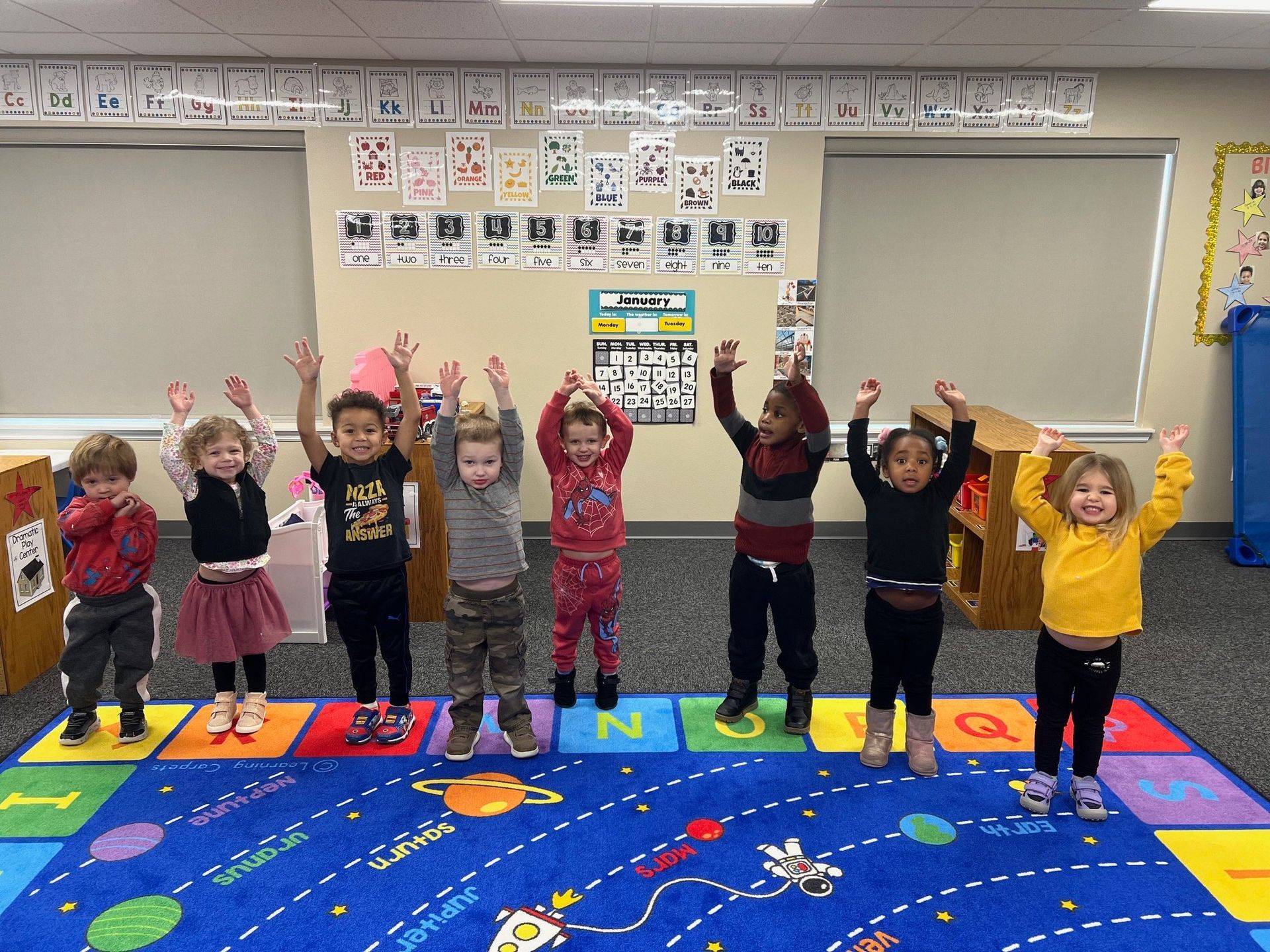 A group of children are standing in a classroom with their hands in the air.