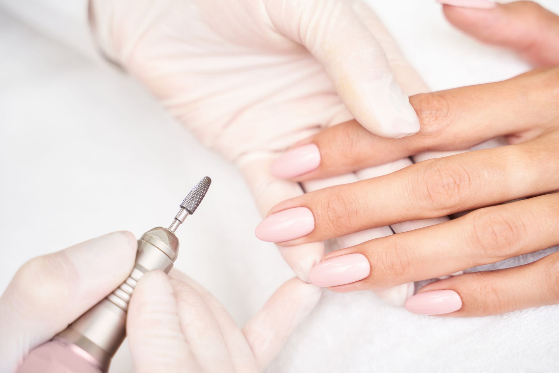 La manicurista usa un torno de uñas en la mano de una clienta, preparándola para la manicura. Ambas manos llevan guantes, en un ambiente luminoso.