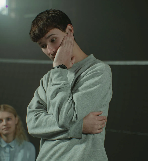 A young man in a grey sweatshirt looks thoughtful, resting his face on his hand, in a classroom.