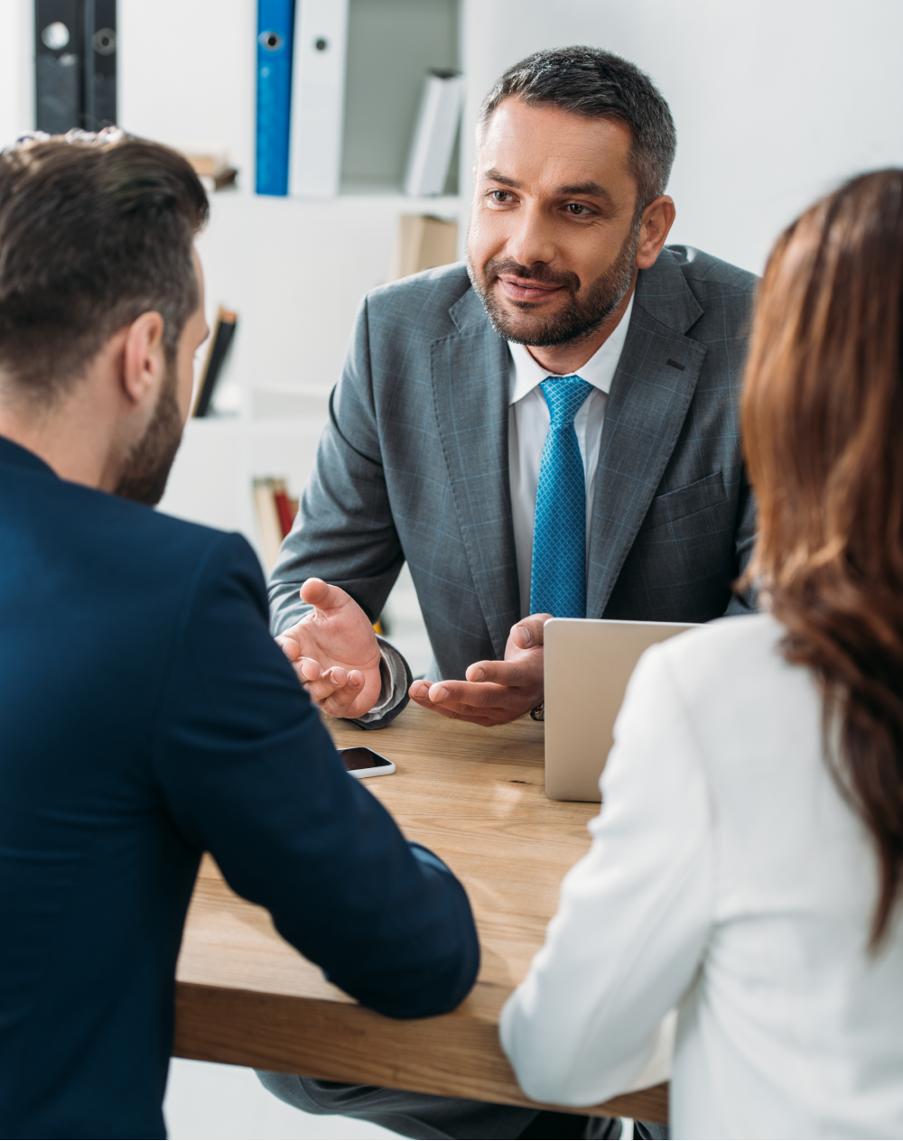 A businessman in a suit gestures while speaking to a couple at a table, in an office setting.
