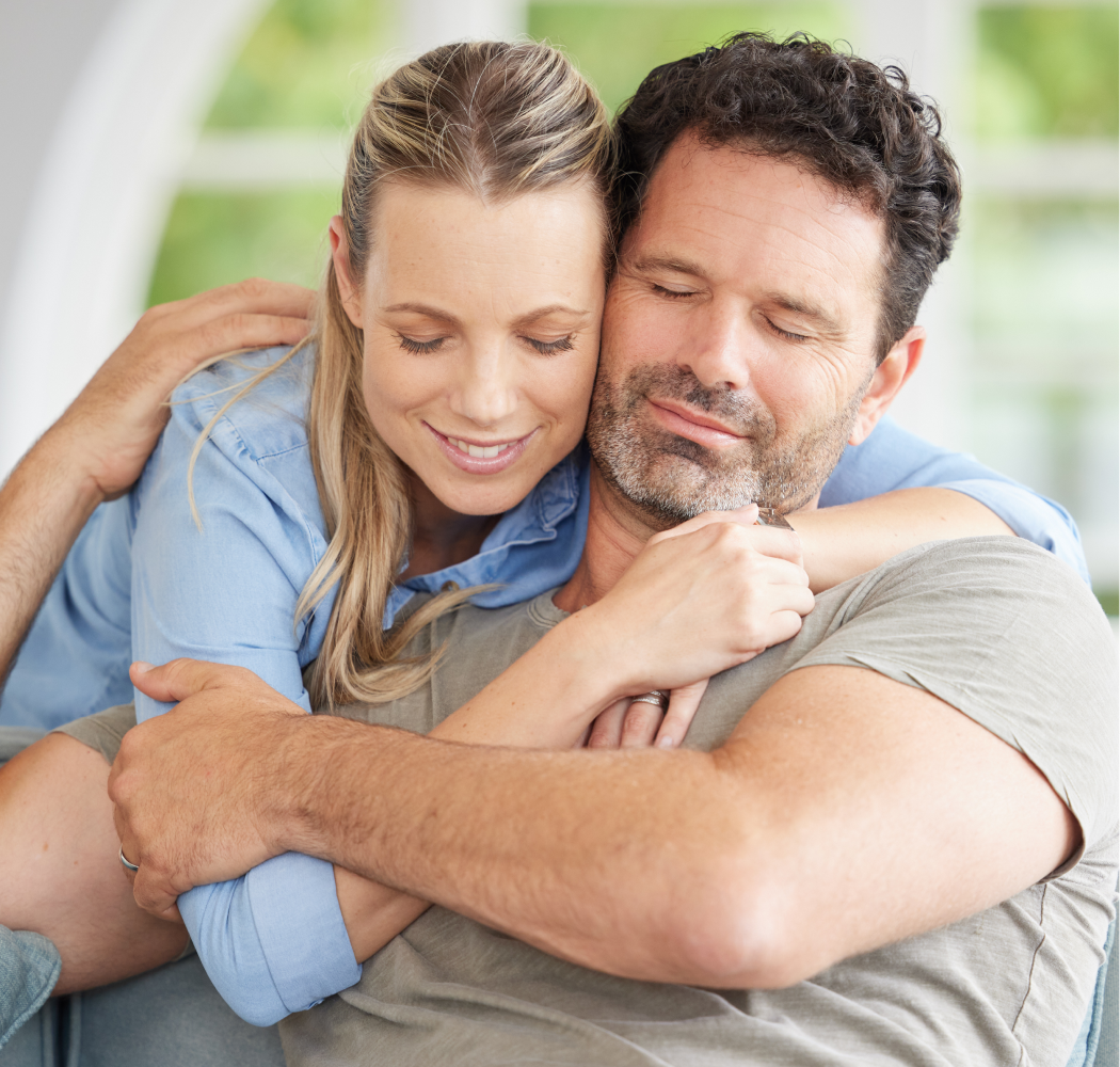 Woman hugging a man, both with eyes closed, smiling, indoors.