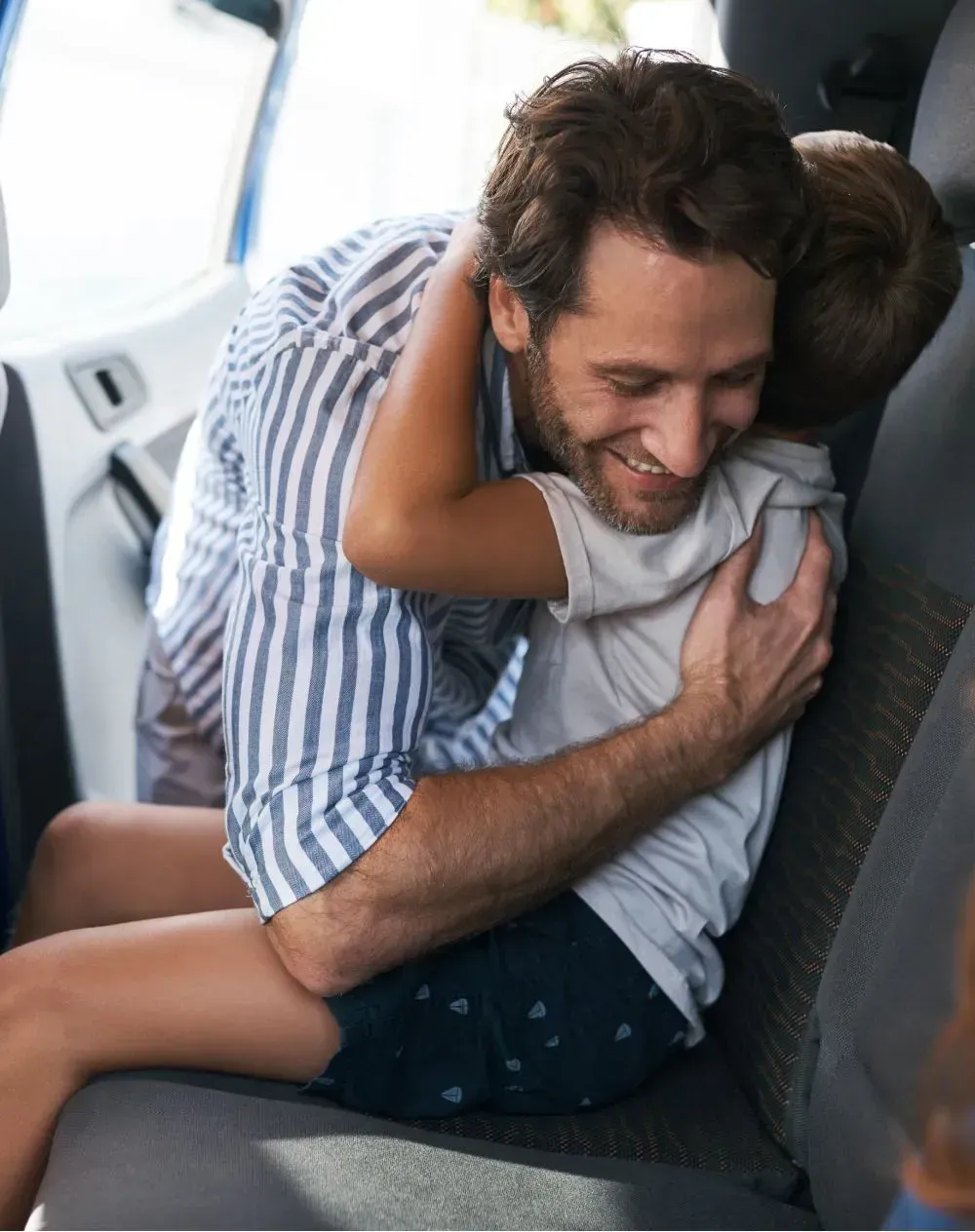 Father and child embrace in a car. The father smiles, wearing a striped shirt.