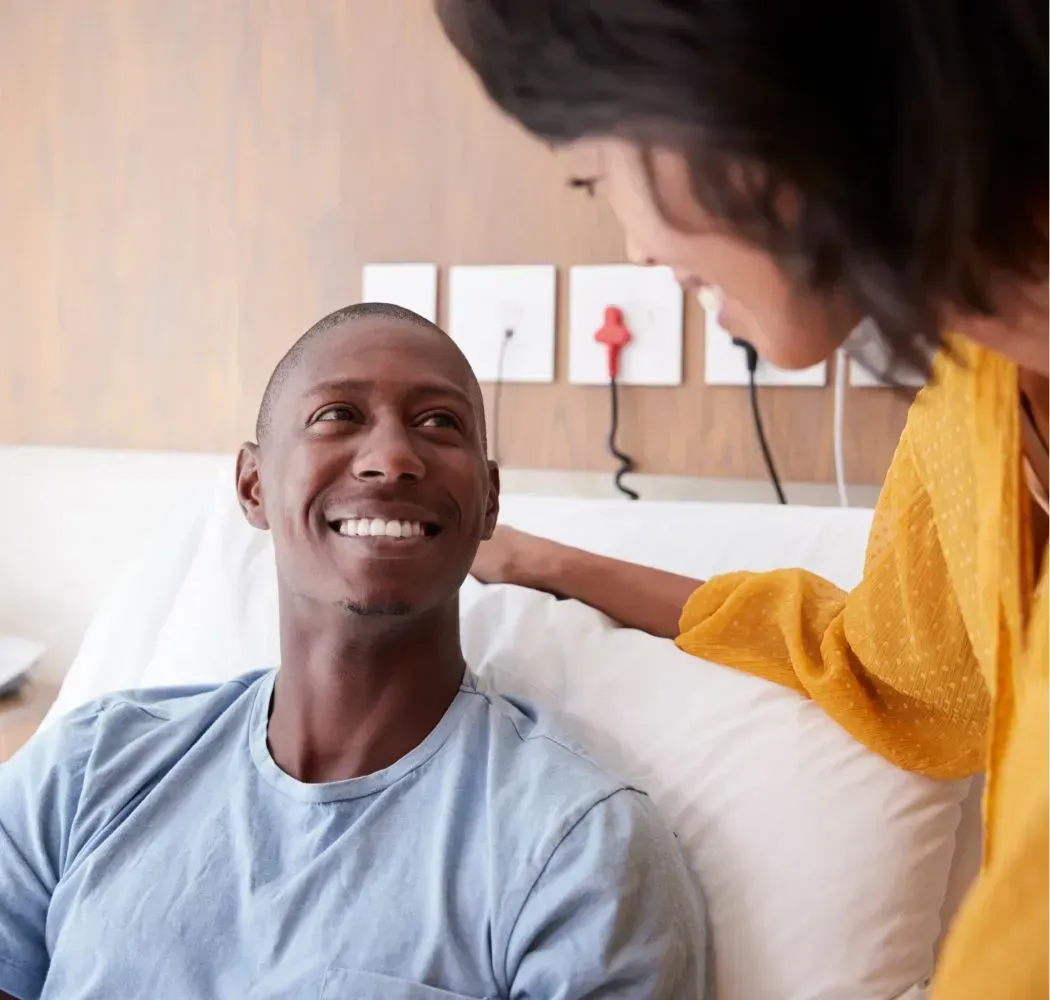 Man in hospital bed smiles at woman, who touches his shoulder.