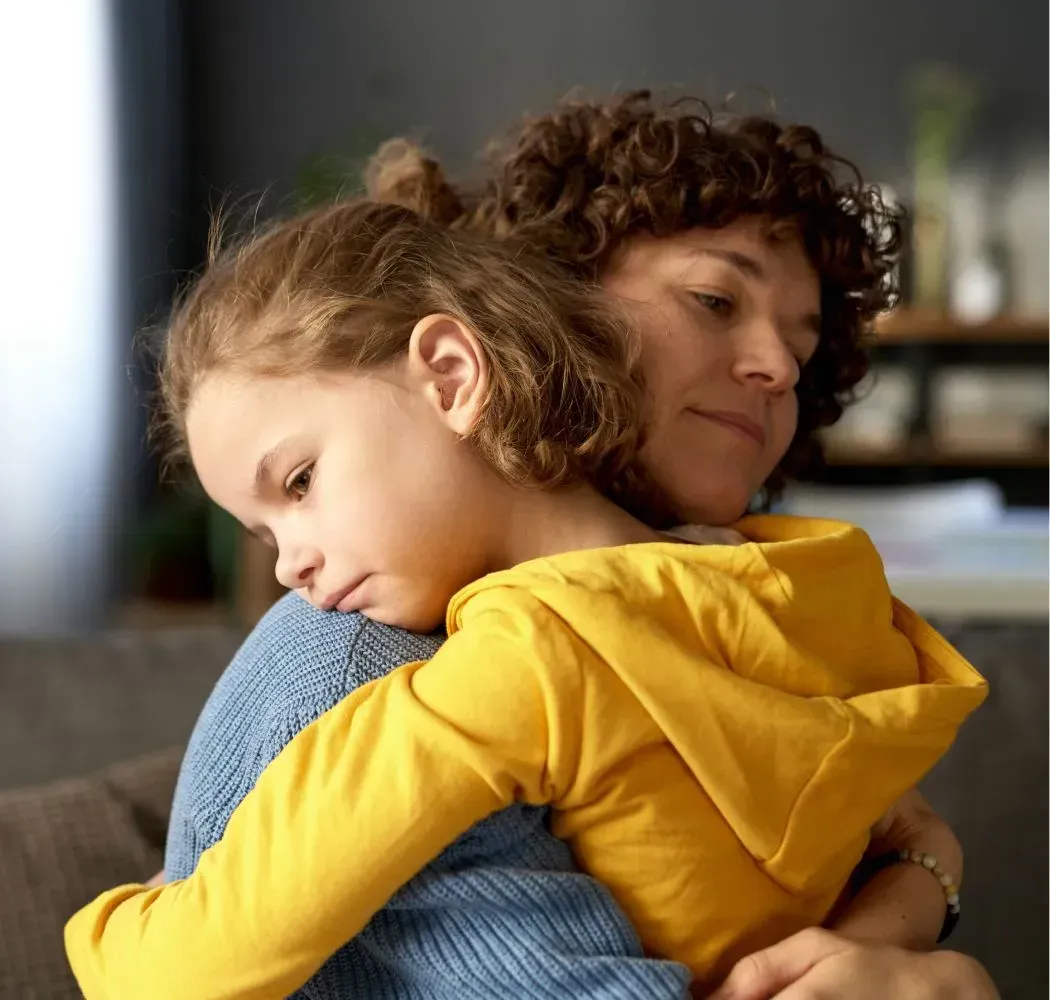 Woman hugging a sad child, indoors. Child wears yellow hoodie, woman, blue sweater.