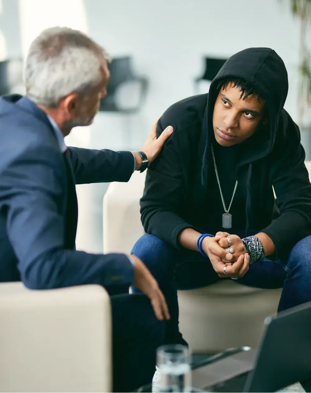 Man in hoodie listens to a man in a suit who has his hand on his shoulder.