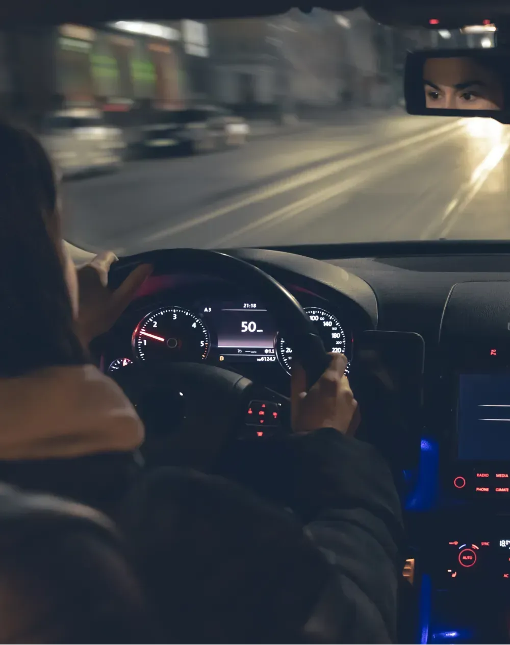 Person driving car at night, illuminated dashboard, city street in background.