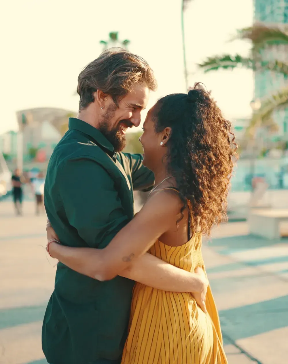 Man and woman embracing, smiling at each other outdoors. Sunny, coastal setting.