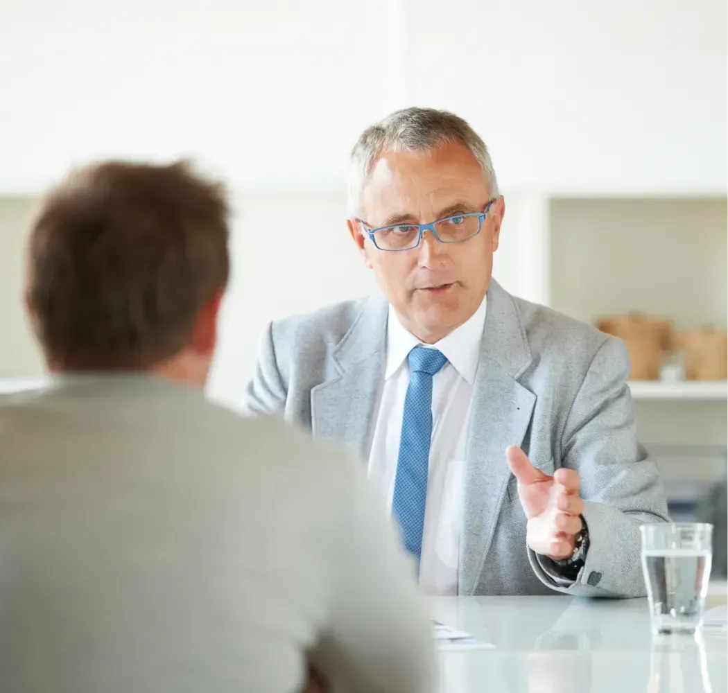 Older man in a suit and tie speaking to a person at a table. A glass of water sits nearby.