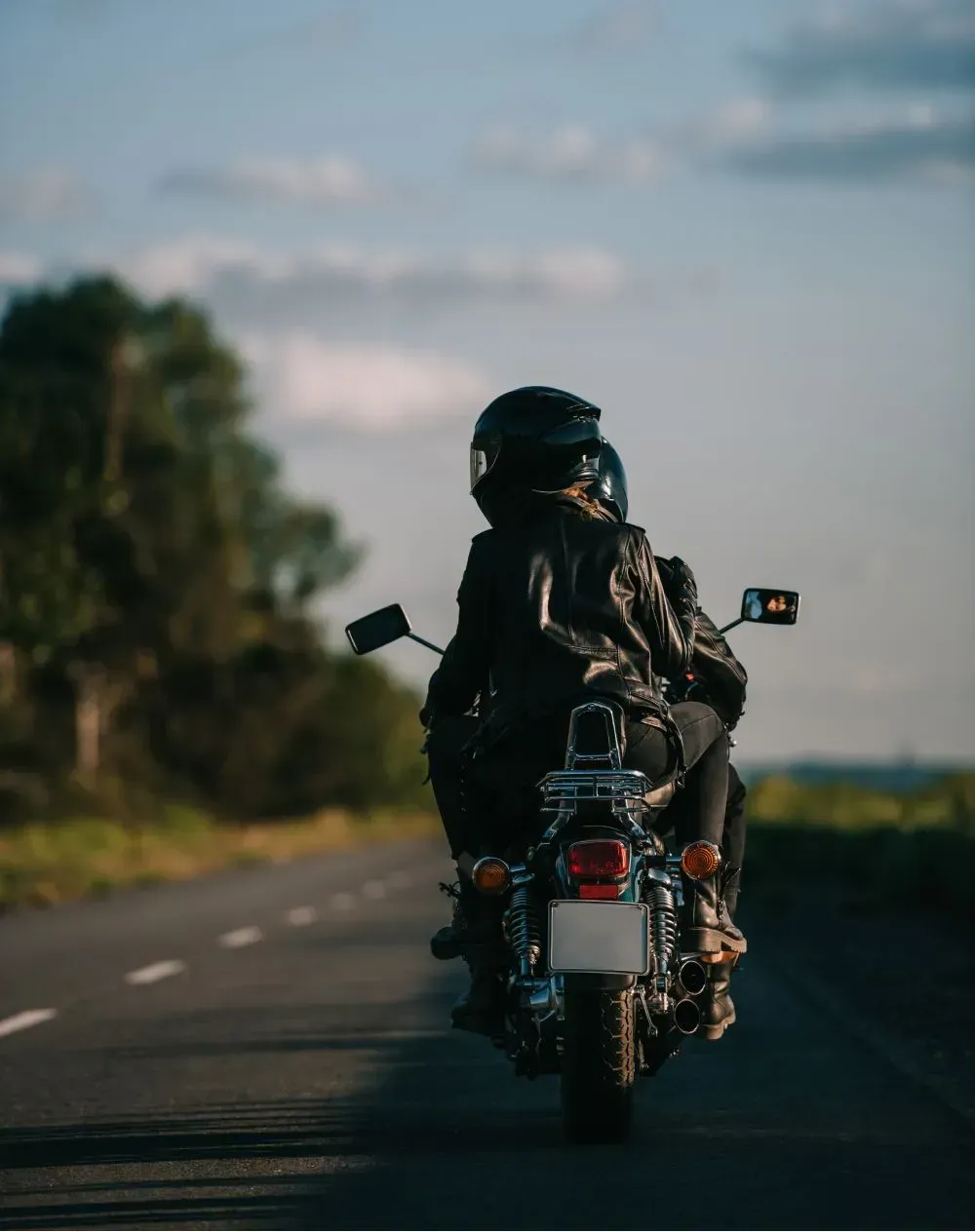 Person on motorcycle rides down a road; wearing a helmet and black leather jacket. Sunny day.