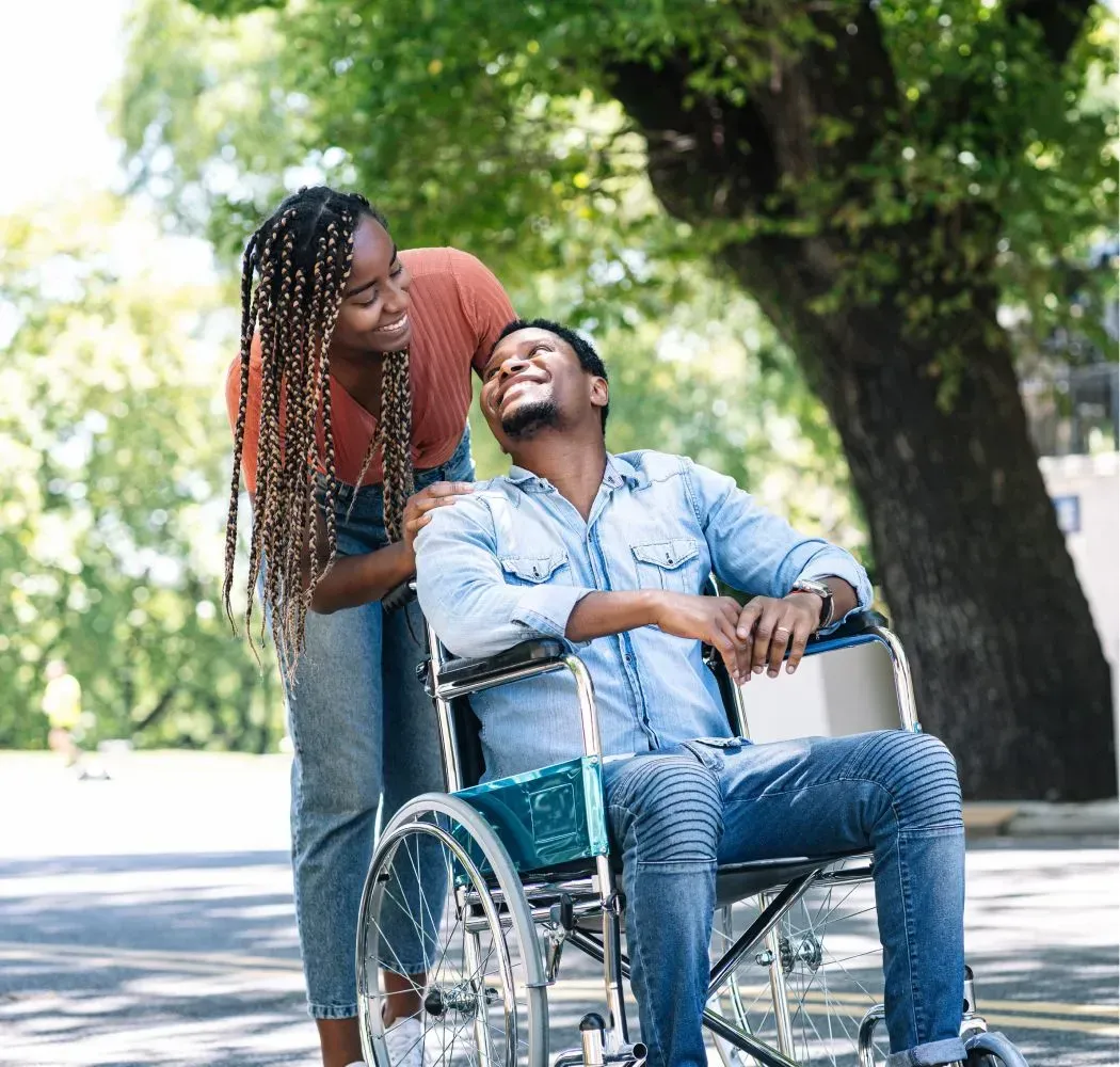 Woman smiles, assisting a man in a wheelchair outdoors. They're both Black, looking at each other happily near a large tree.