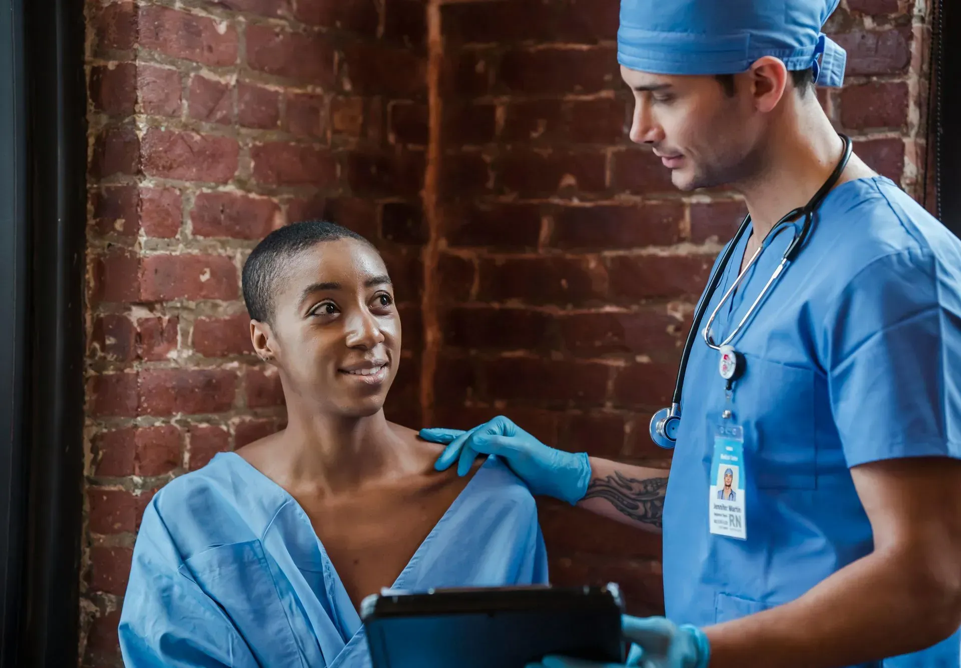 Doctor in blue scrubs, touches shoulder of Black female patient in blue gown, smiles, in a brick room.