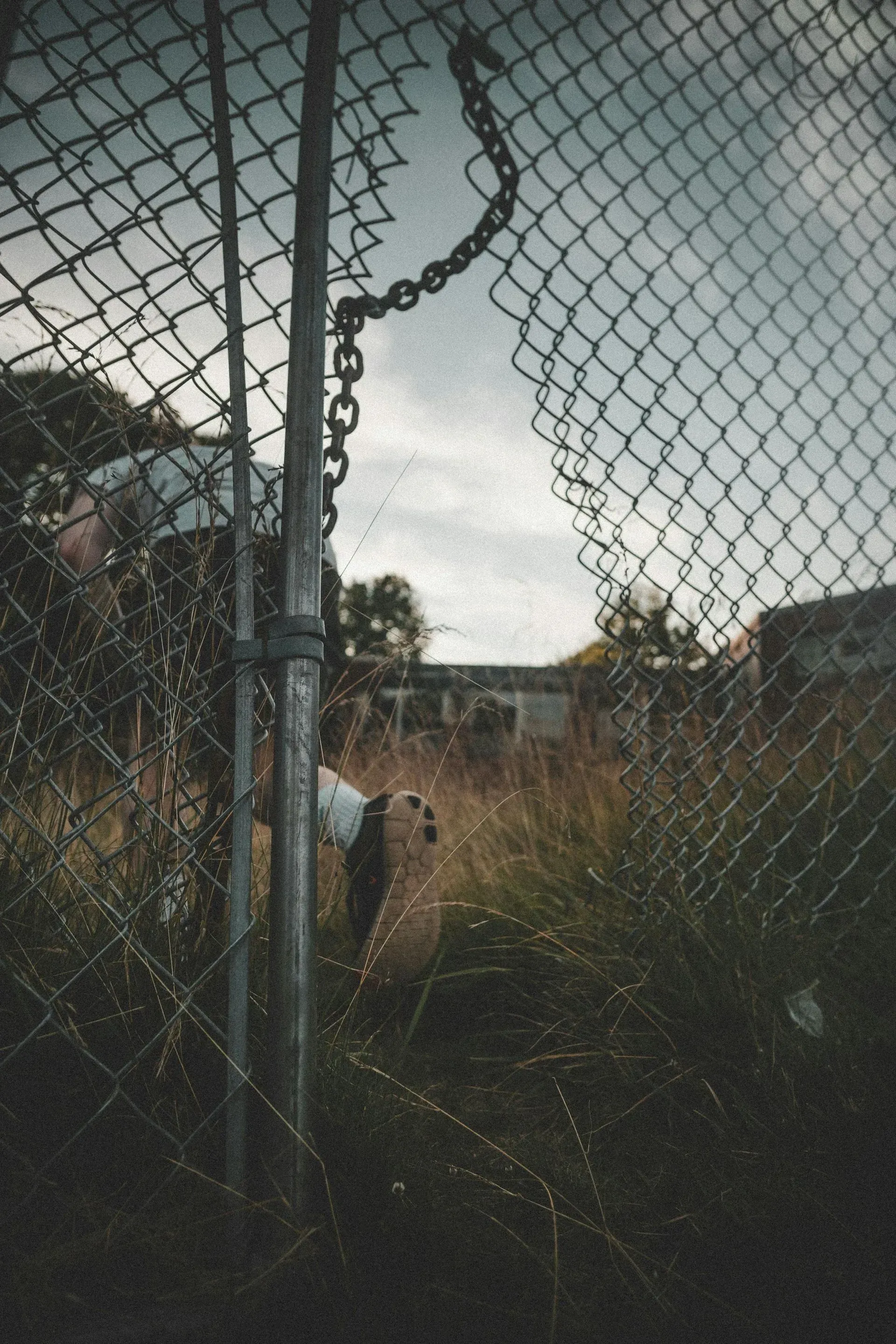 Chain-link fence with a hole, revealing overgrown grass and a building under a cloudy sky.