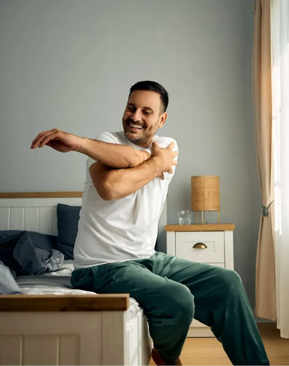 Man stretching his arm on a bed, smiling. Bedroom setting with a bedside table.