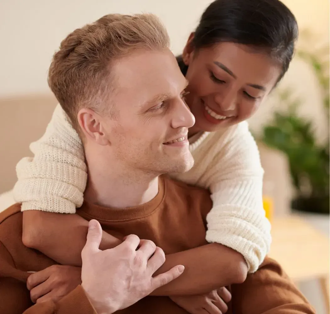 Woman hugs a smiling man from behind. They are indoors and look happy.