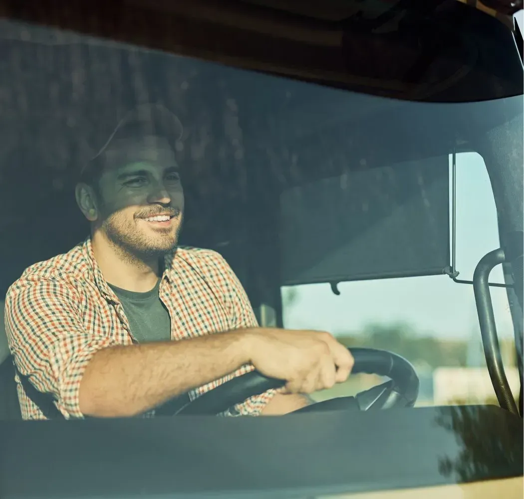 Smiling man driving a truck, wearing plaid shirt. Sunlight visible, sky in background.