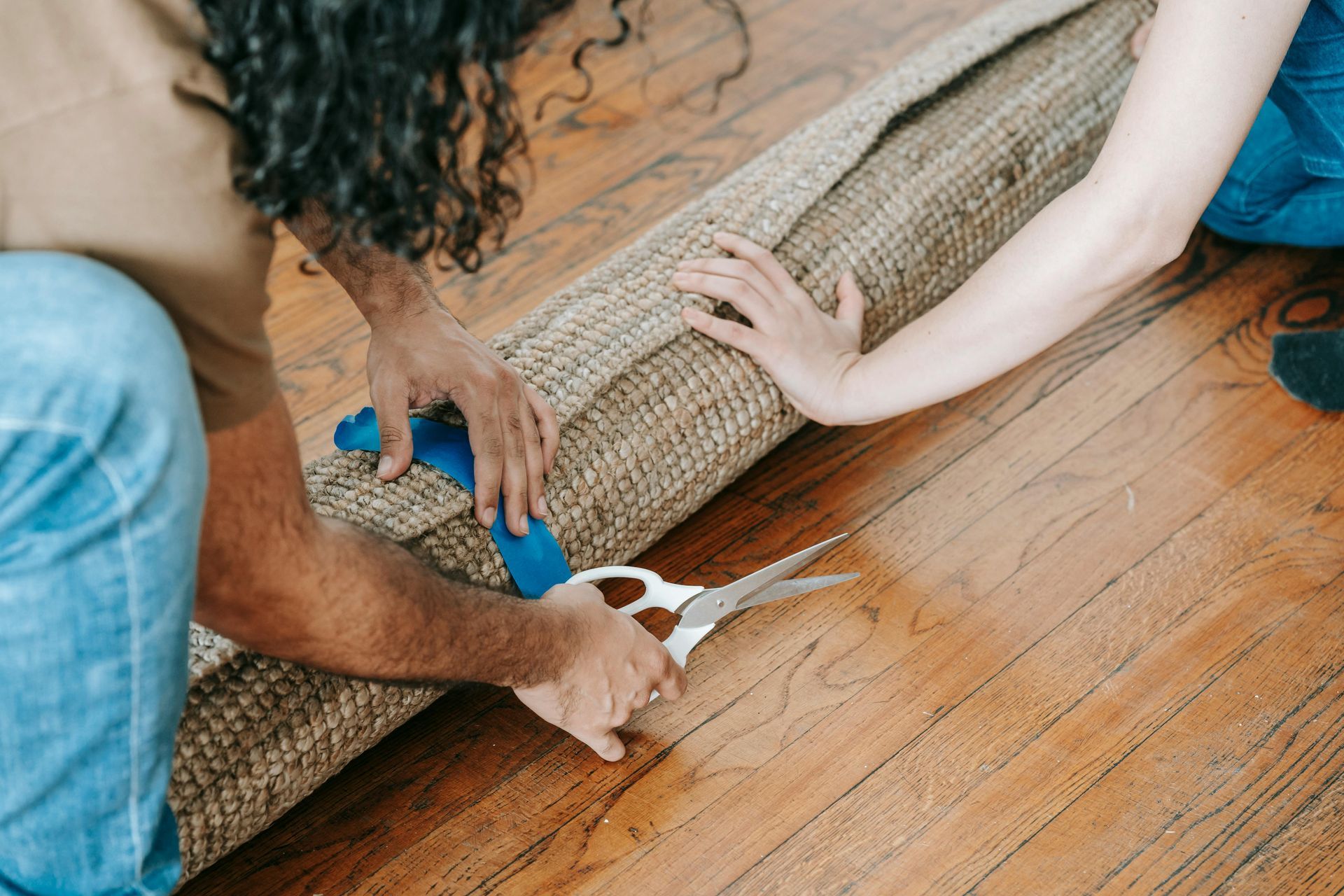 Two people cutting a rolled rug with scissors on a hardwood floor.