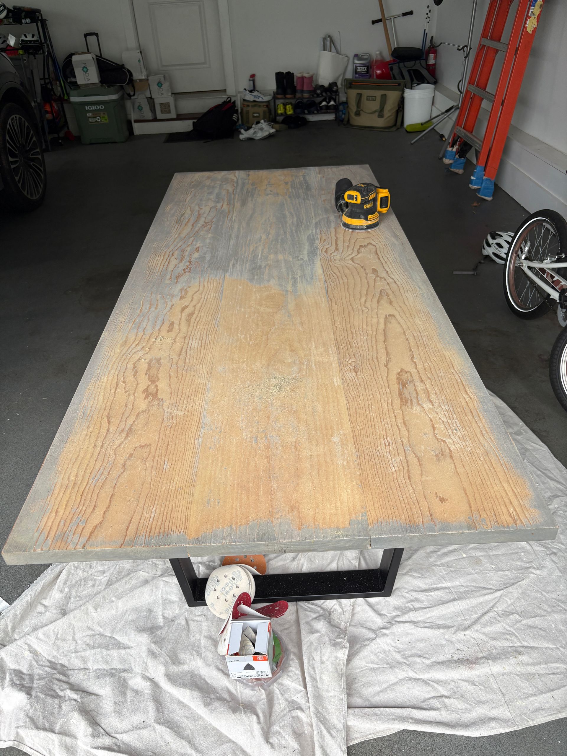 A rectangular wooden table on a white sheet in a garage, with a sander on it and tools below.