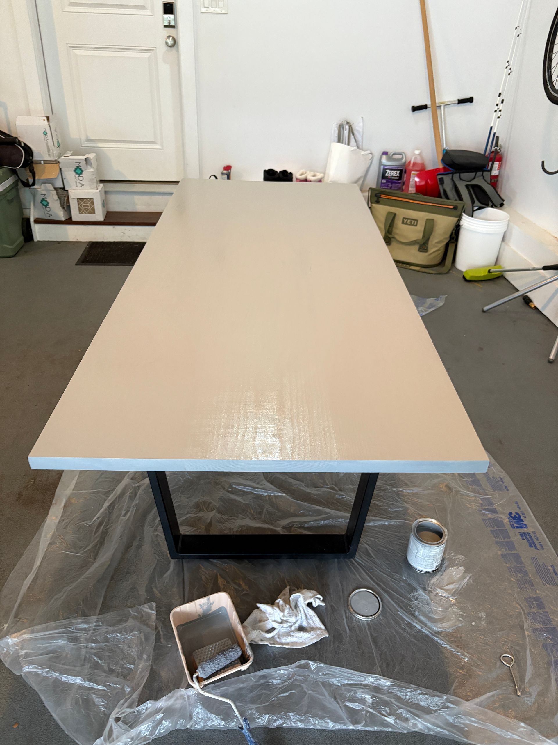 Long rectangular table, painted light gray, on a black metal base, in a garage. Paint supplies visible.