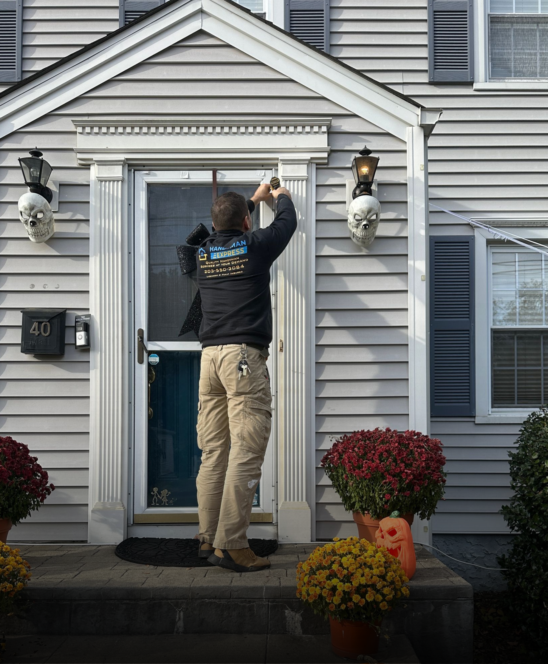 A person in a black long-sleeve shirt and khaki pants stands on a front porch attaching Halloween decor to a door frame.