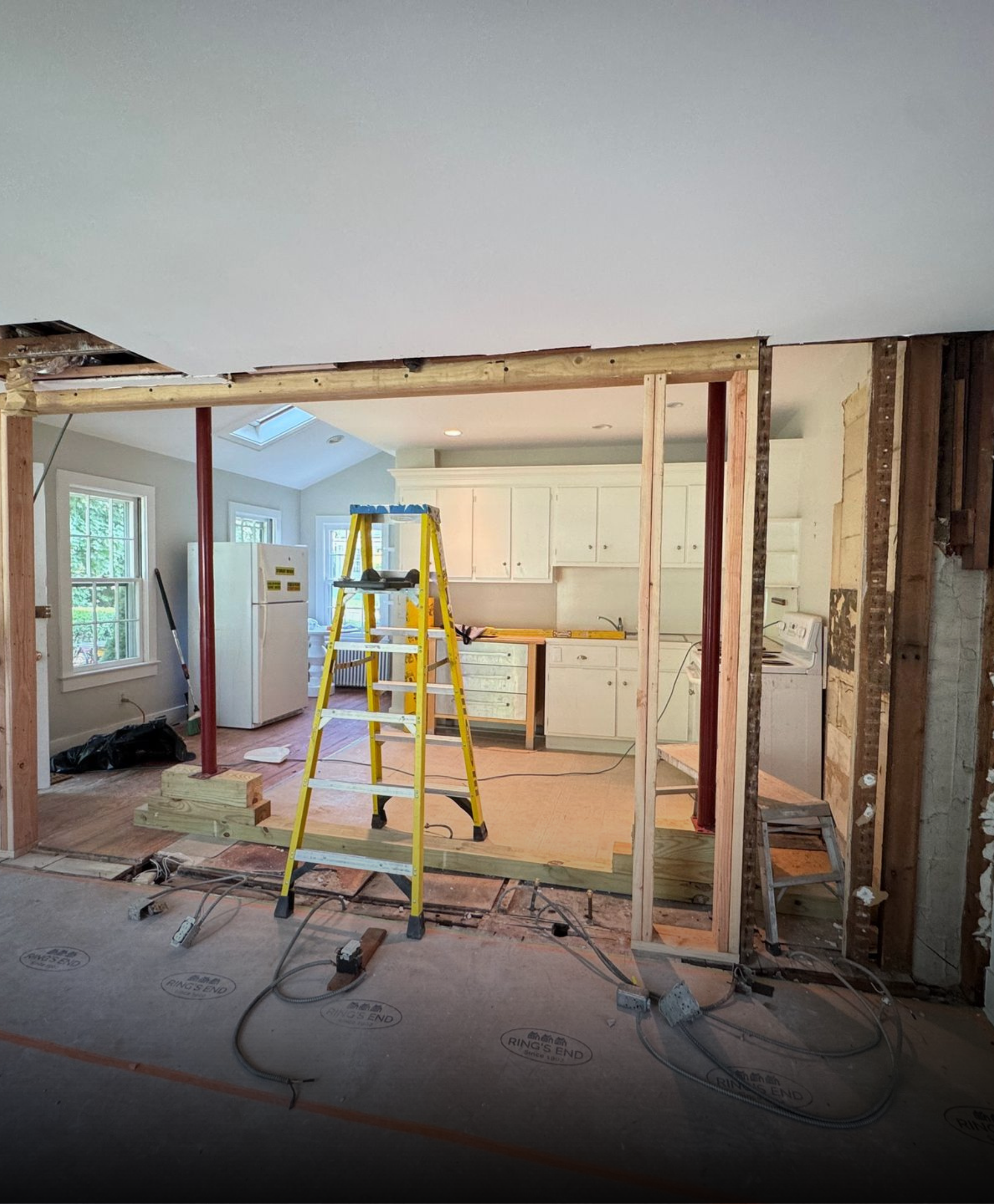 A yellow stepladder stands in a home renovation zone where a wall has been removed, revealing a white kitchen beyond.