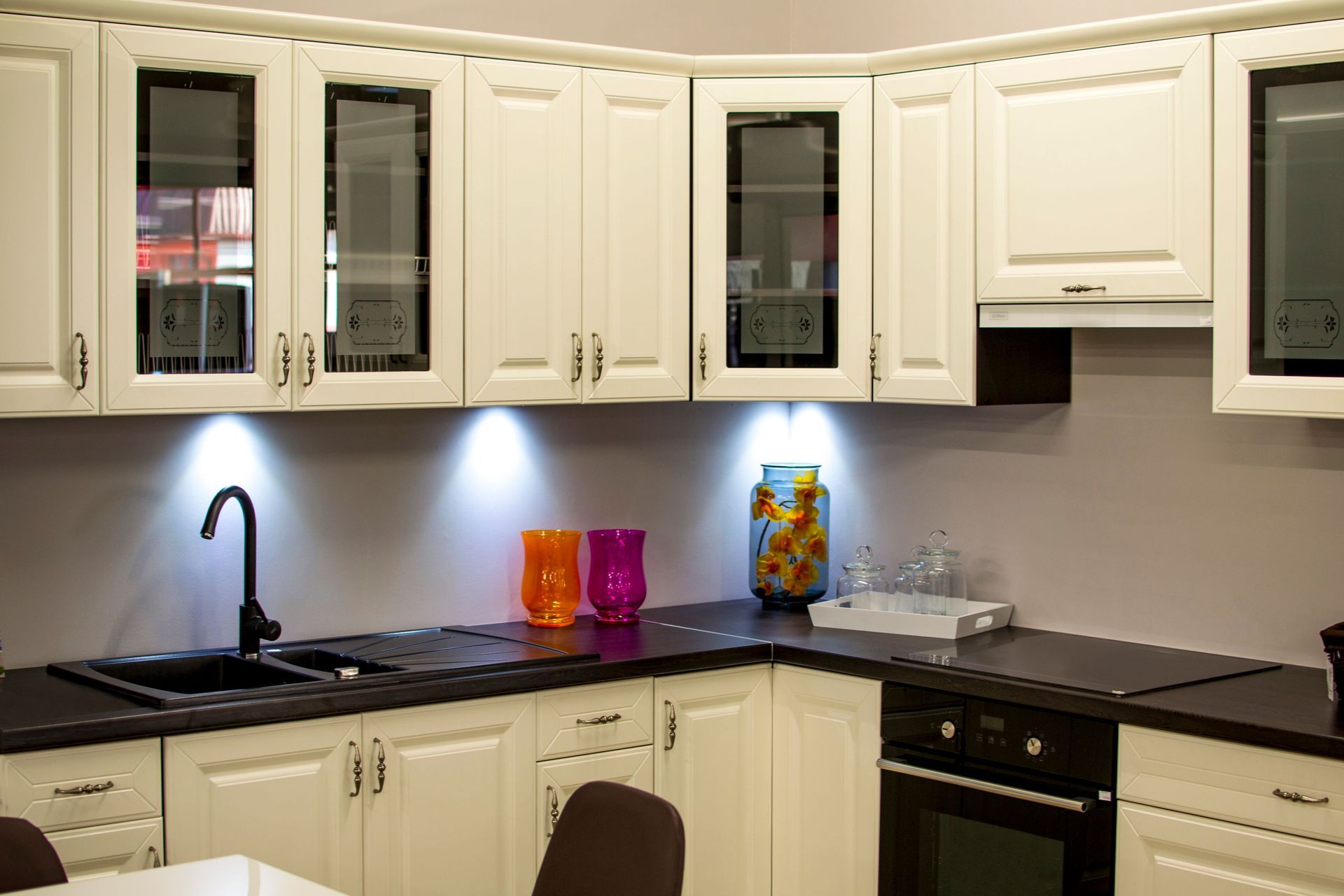 Modern white kitchen with black countertops, glass-front cabinets, and a lit faucet over the sink.