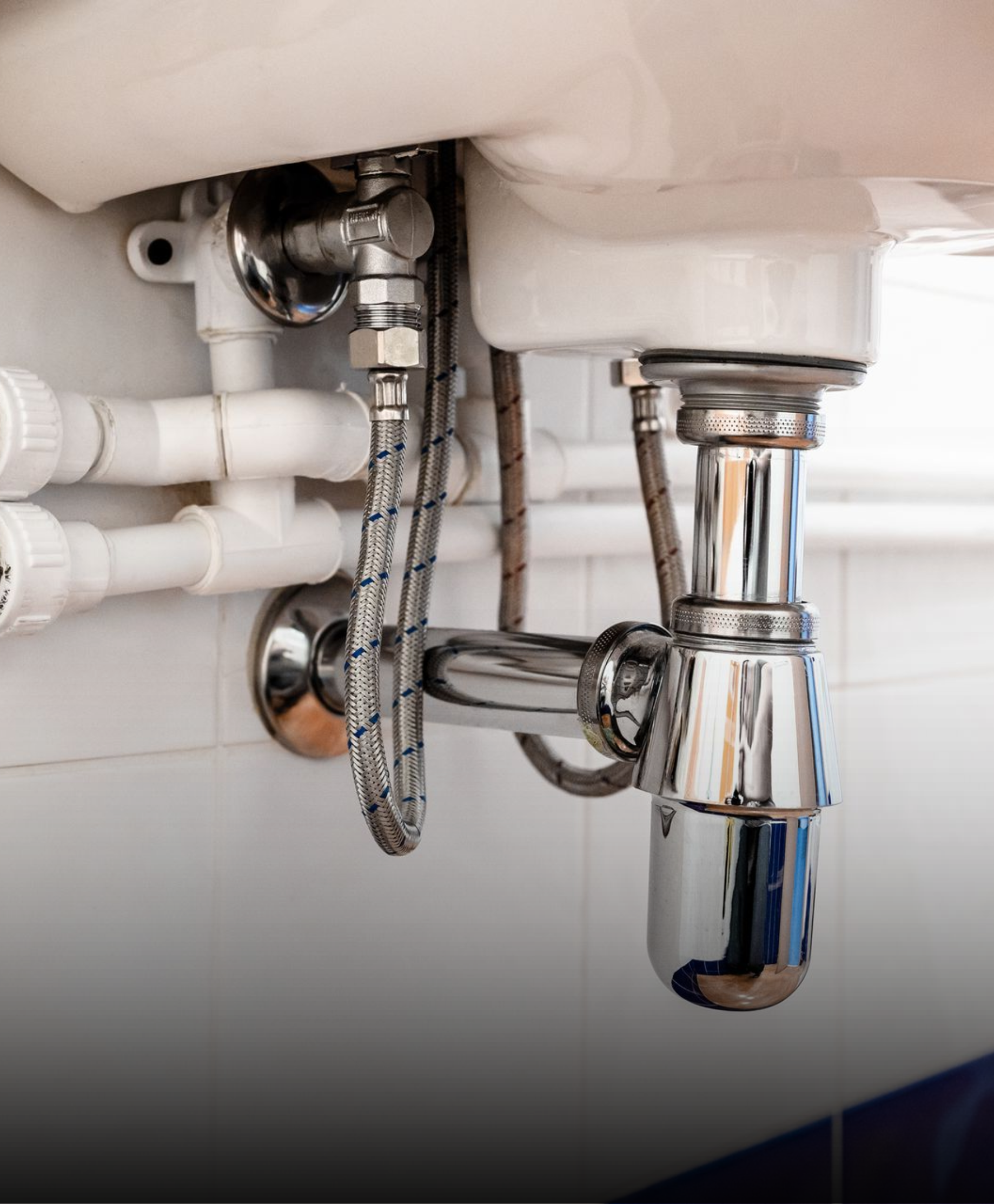 A chrome bottle trap and flexible water supply hoses connected beneath a bathroom sink against a white tiled wall.