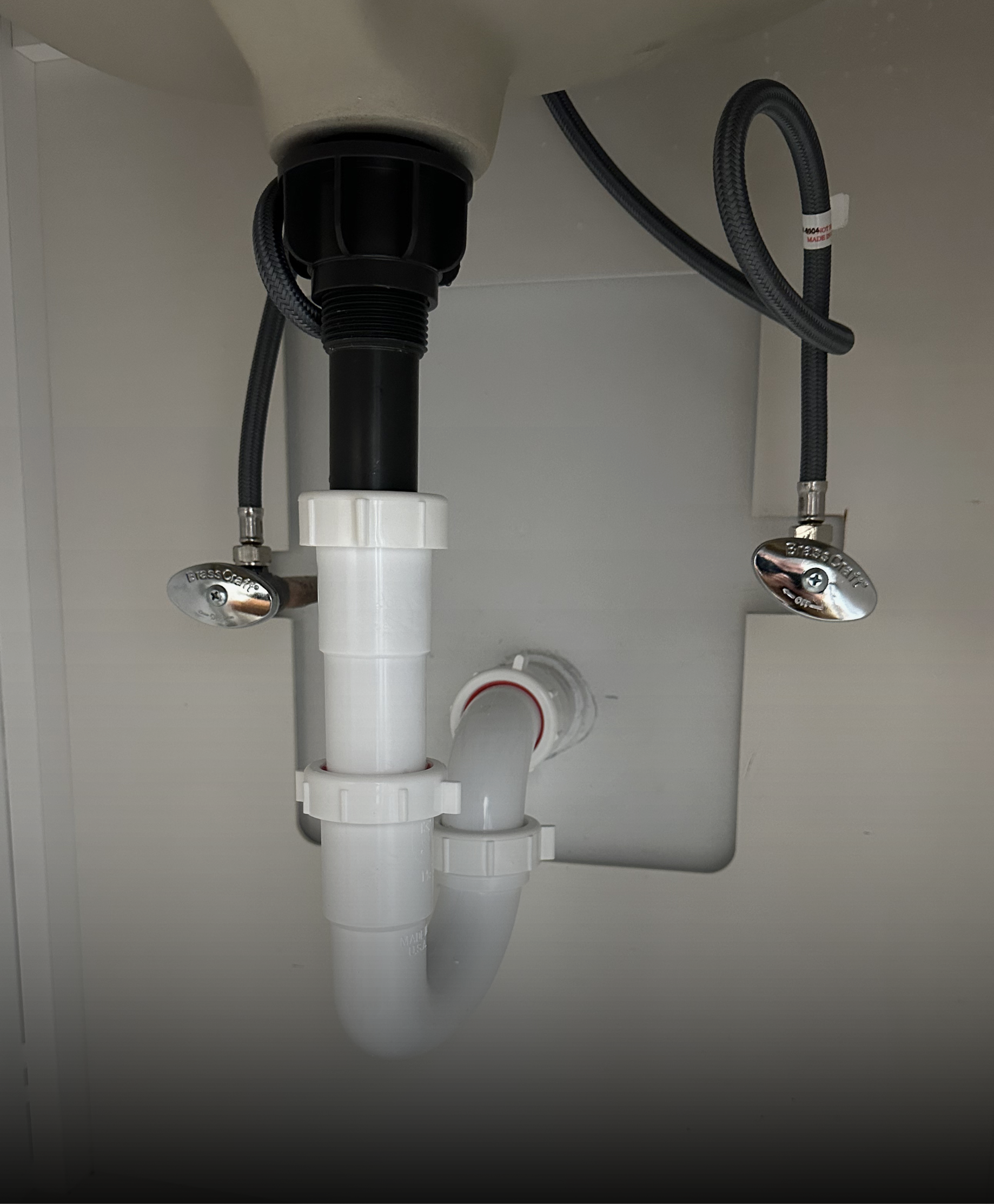 A chrome bottle trap and flexible water supply hoses connected beneath a bathroom sink against a white tiled wall.